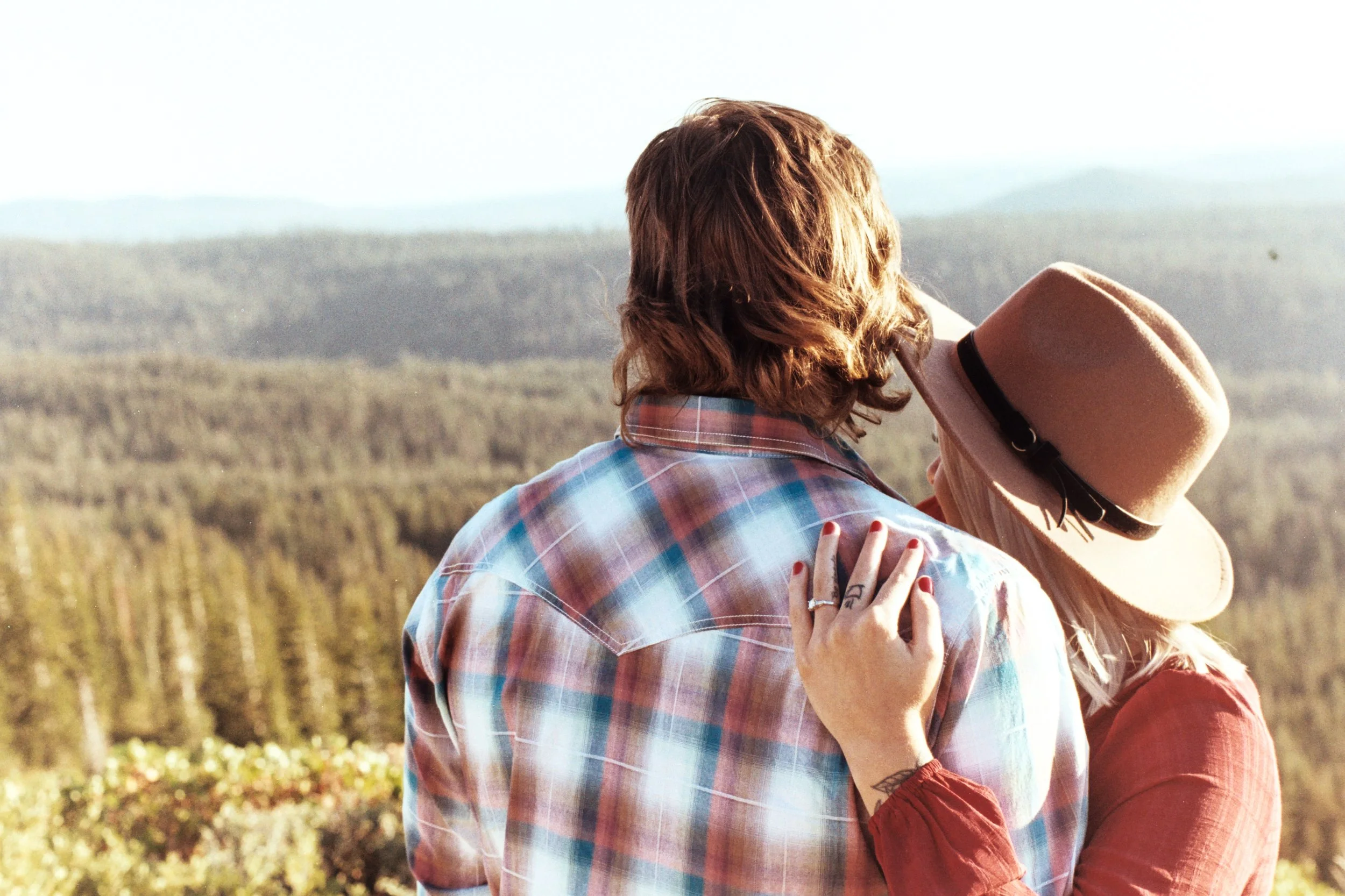 Two women embrace outdoors in a nature setting during daytime, one wearing a plaid shirt and the other a hat, with a blurred landscape of fields and hills in the background.