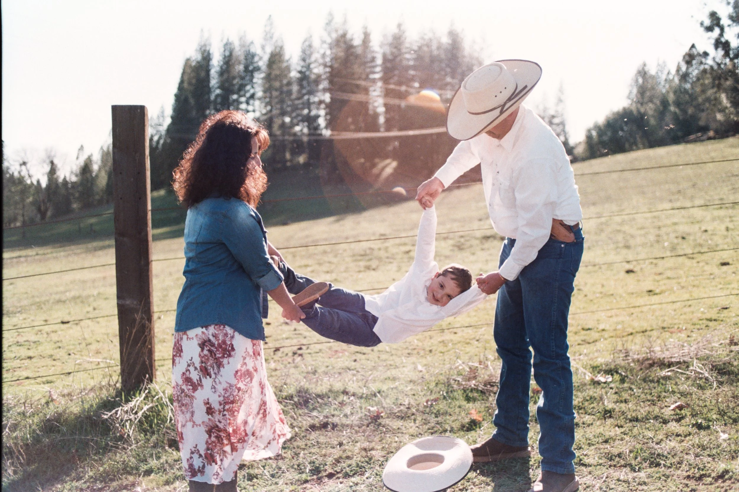 Three people holding a child by the arms in a grassy field, with trees and a wooden fence in the background on a sunny day.