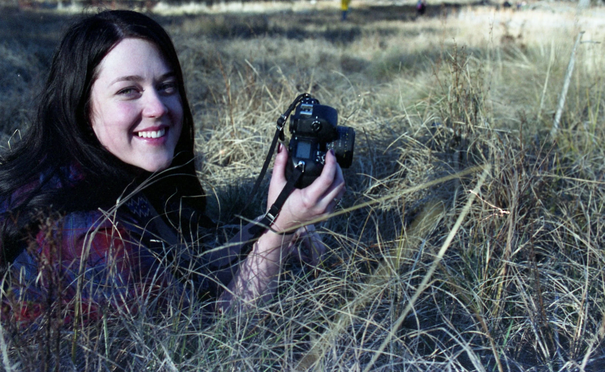 A woman with dark hair smiling while holding a camera in a field of dry grass.