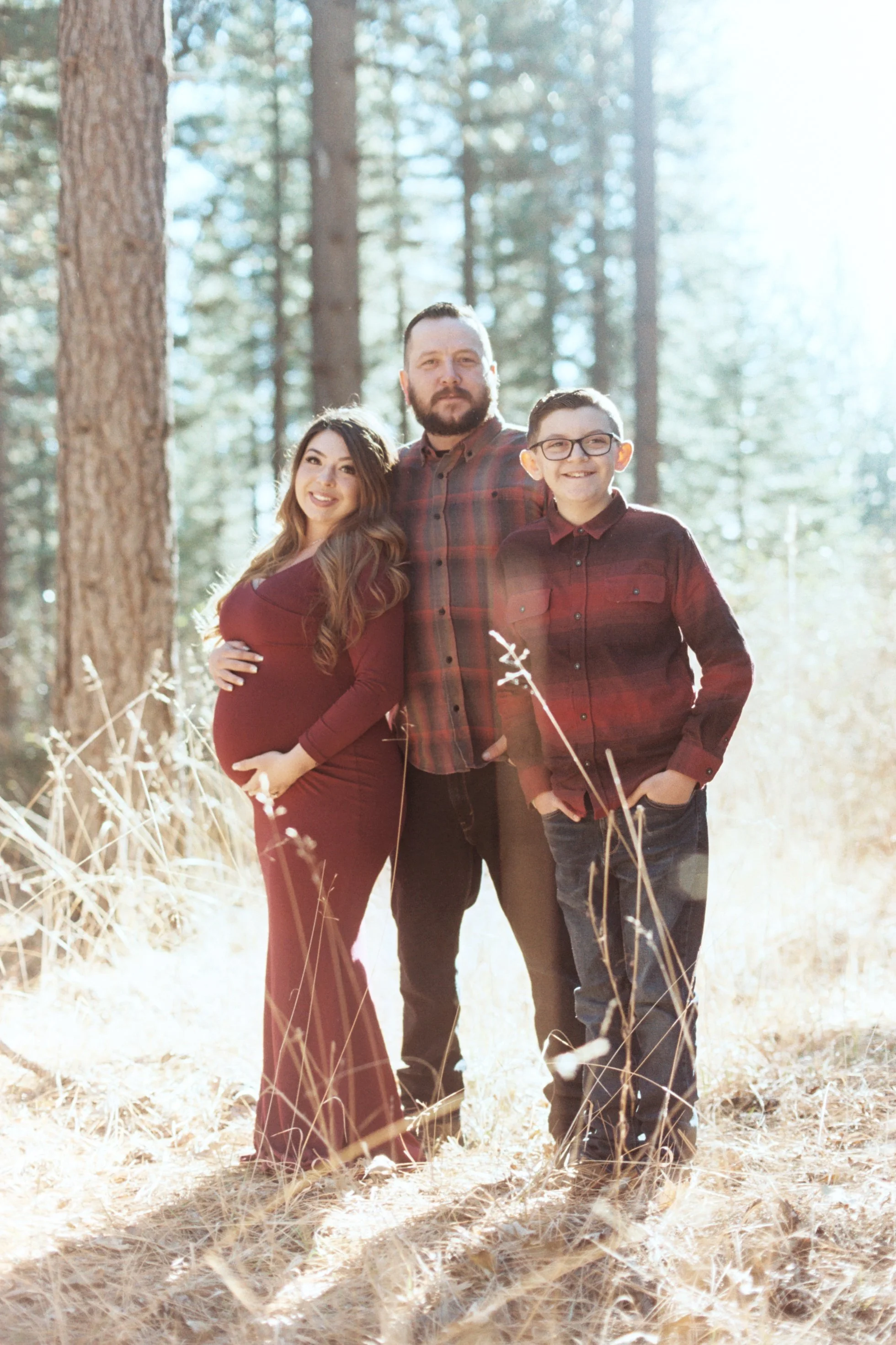 A family of three standing in a sunlit forest, with a pregnant woman on the left, a man in the middle, and a young boy on the right, all smiling and dressed in matching burgundy and plaid shirts.