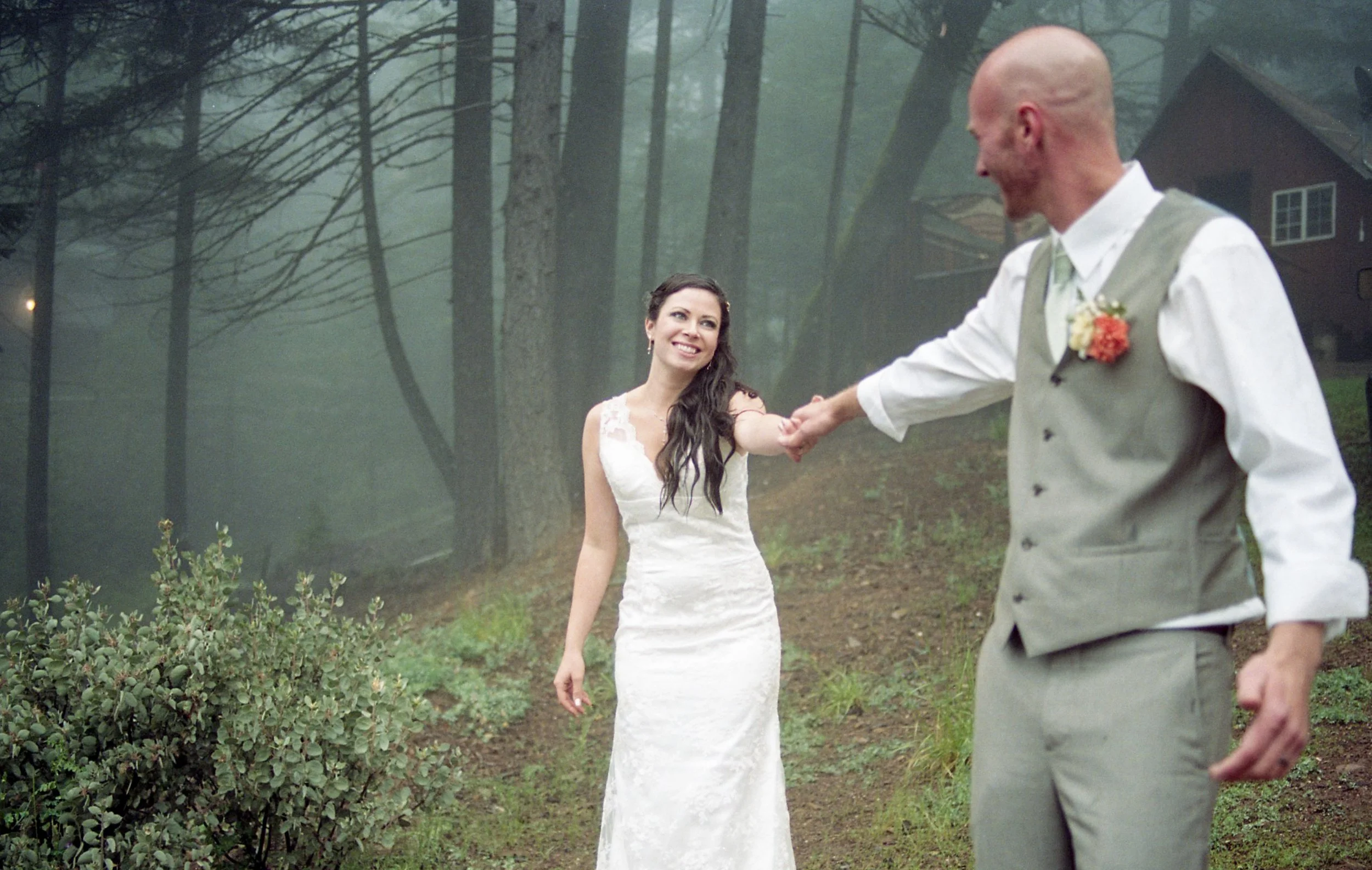 Bride and groom holding hands and smiling in a misty outdoor setting with trees and a house in the background.