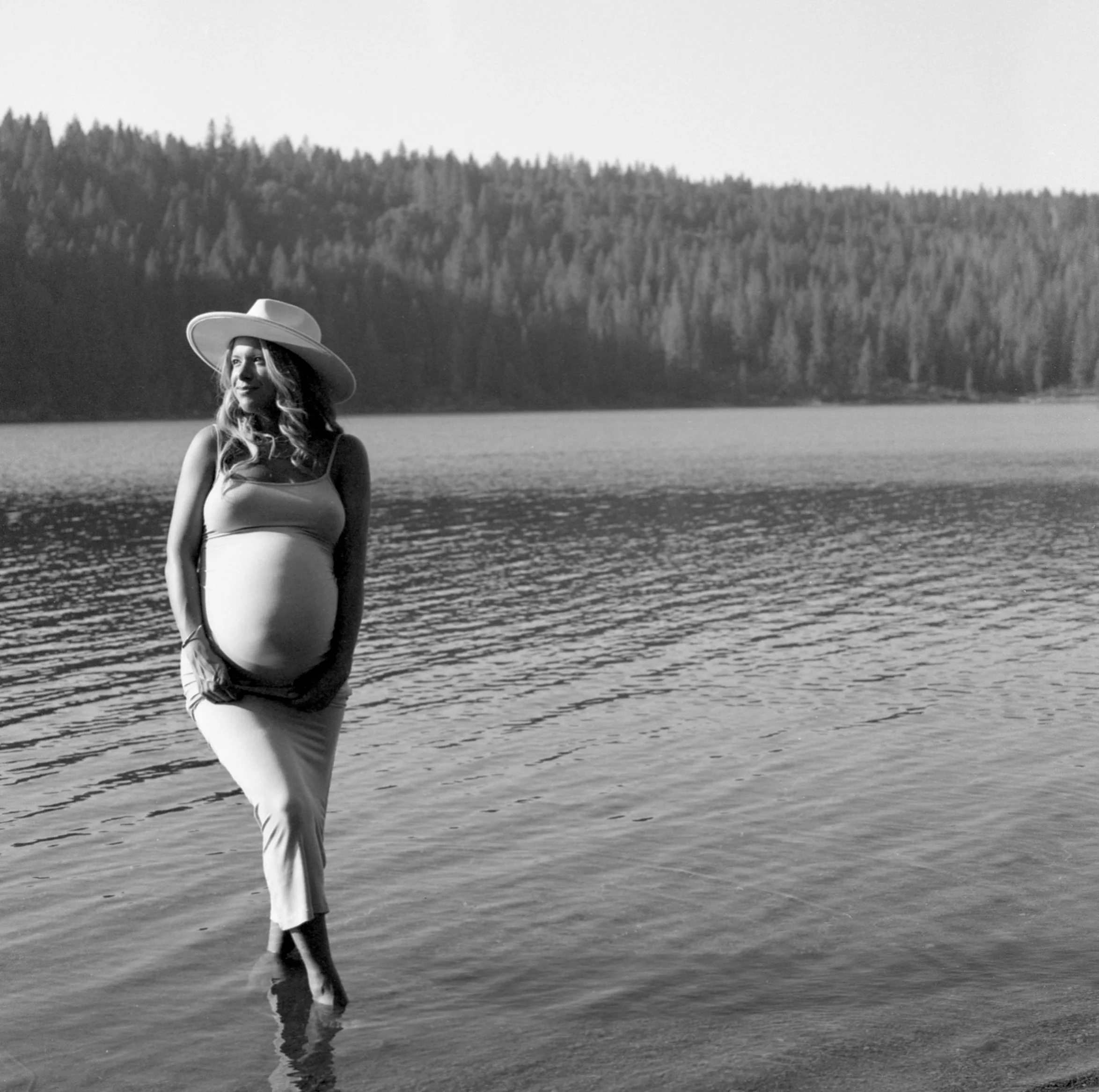 A woman wearing a wide-brimmed hat and sleeveless top standing in shallow water at a lake or river, with a forested hill or mountain in the background.