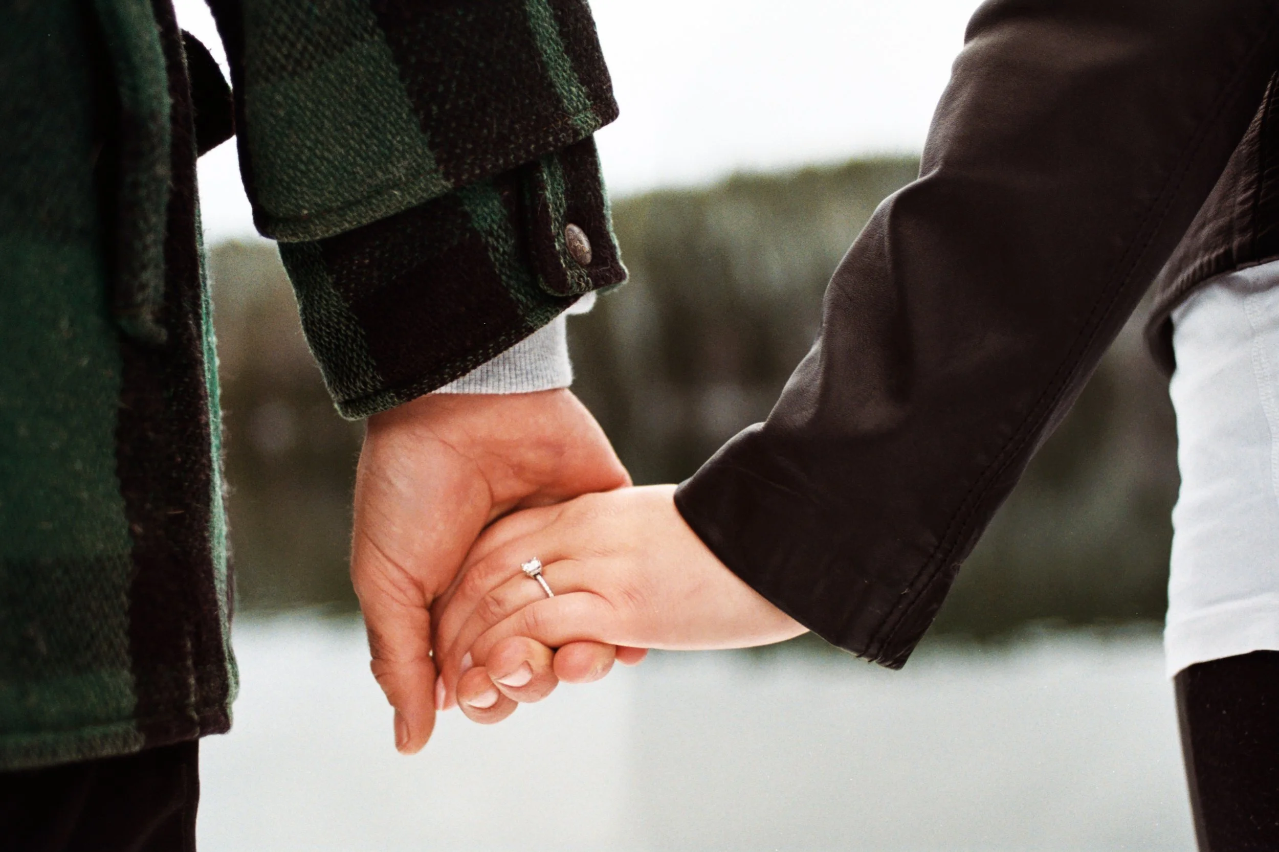 A close-up of a couple holding hands, with the woman's hand showing an engagement ring, while they hold hands gently, signifying a romantic or committed relationship.