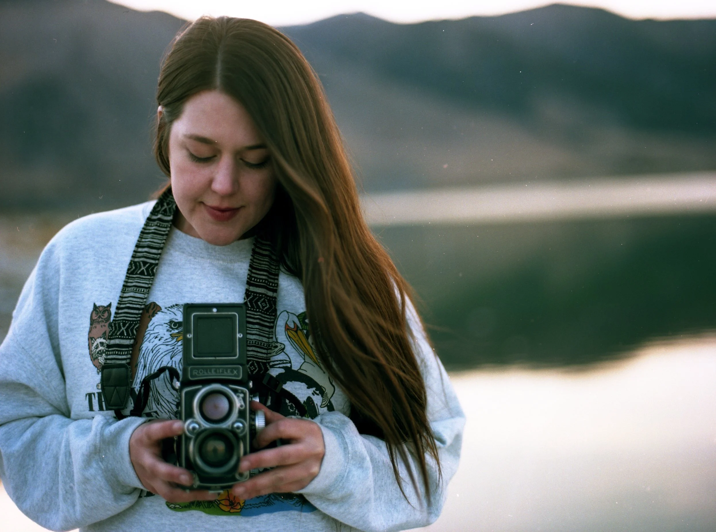 A young woman with long red hair standing outdoors near a body of water, looking down at a vintage Rolleiflex camera she is holding.