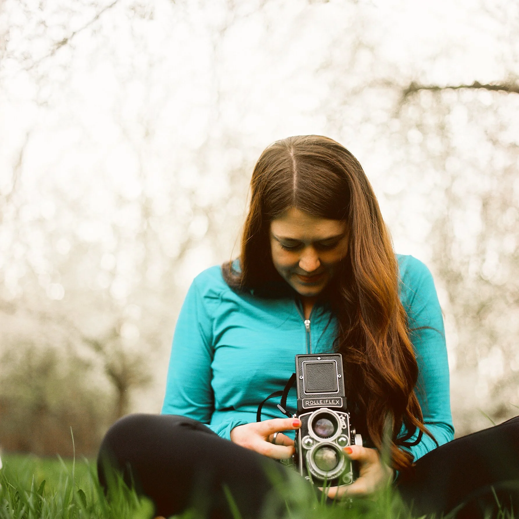 Woman with long brown hair wearing a blue jacket, holding a vintage camera, sitting on the grass outdoors, with trees and sunlight in the background.