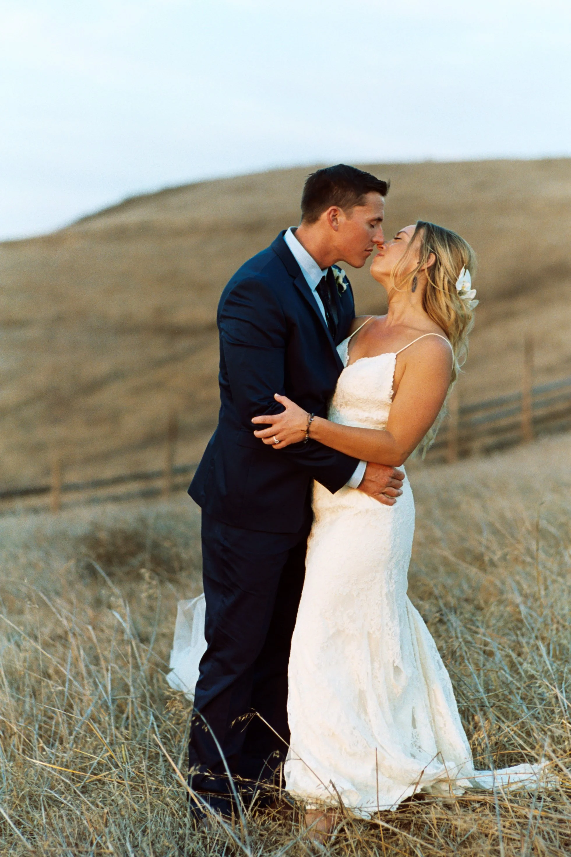 A newlywed couple standing in a dry, grassy field, sharing a romantic moment. The groom, dressed in a navy blue suit, gently holds the bride, who is wearing a white lace wedding dress with spaghetti straps, and has a white flower in her blonde hair.