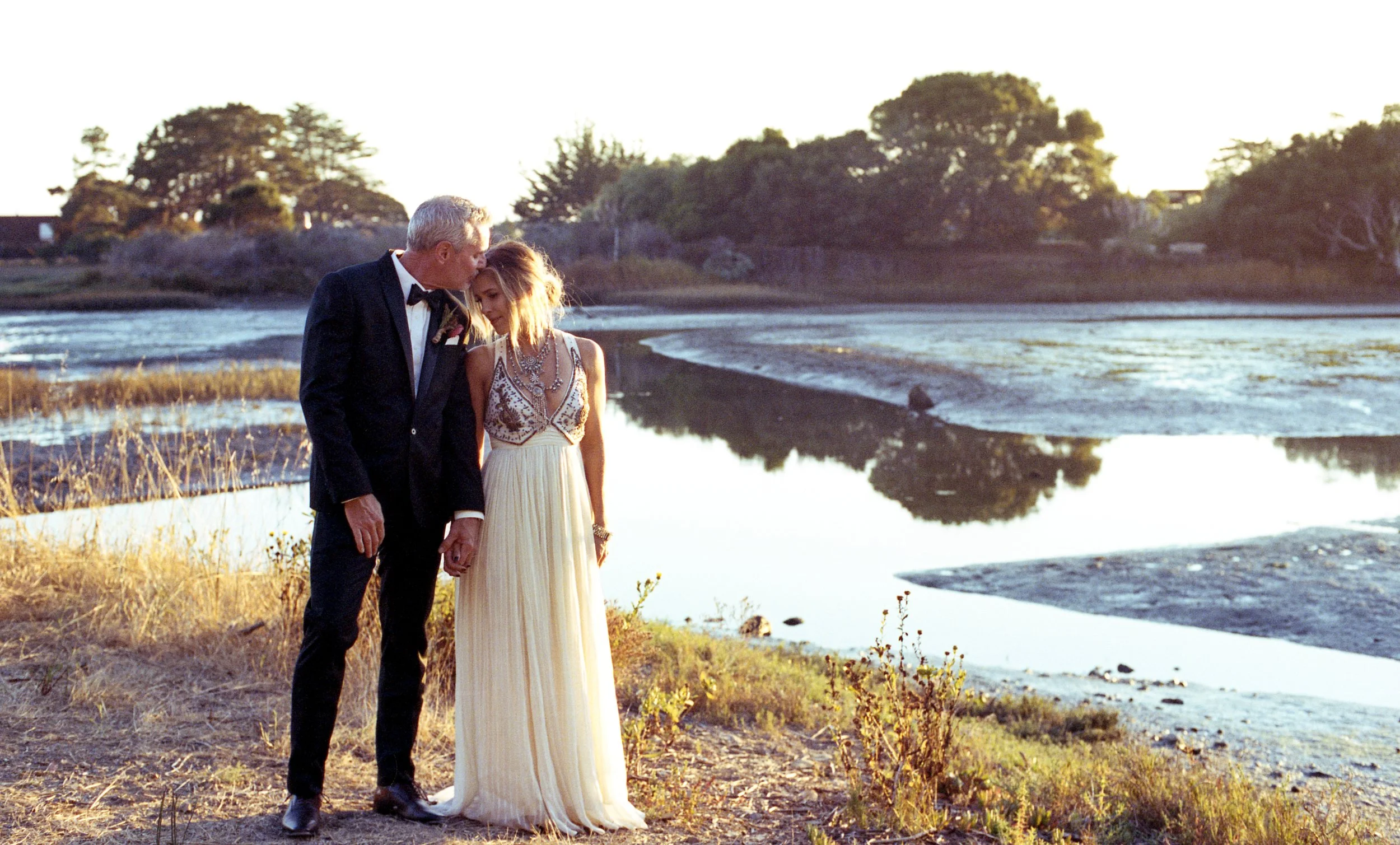 A romantic scene of an older man in a tuxedo kissing a young woman on the forehead, standing by a river during sunset, dressed in a flowing white gown with intricate beading and jewelry.