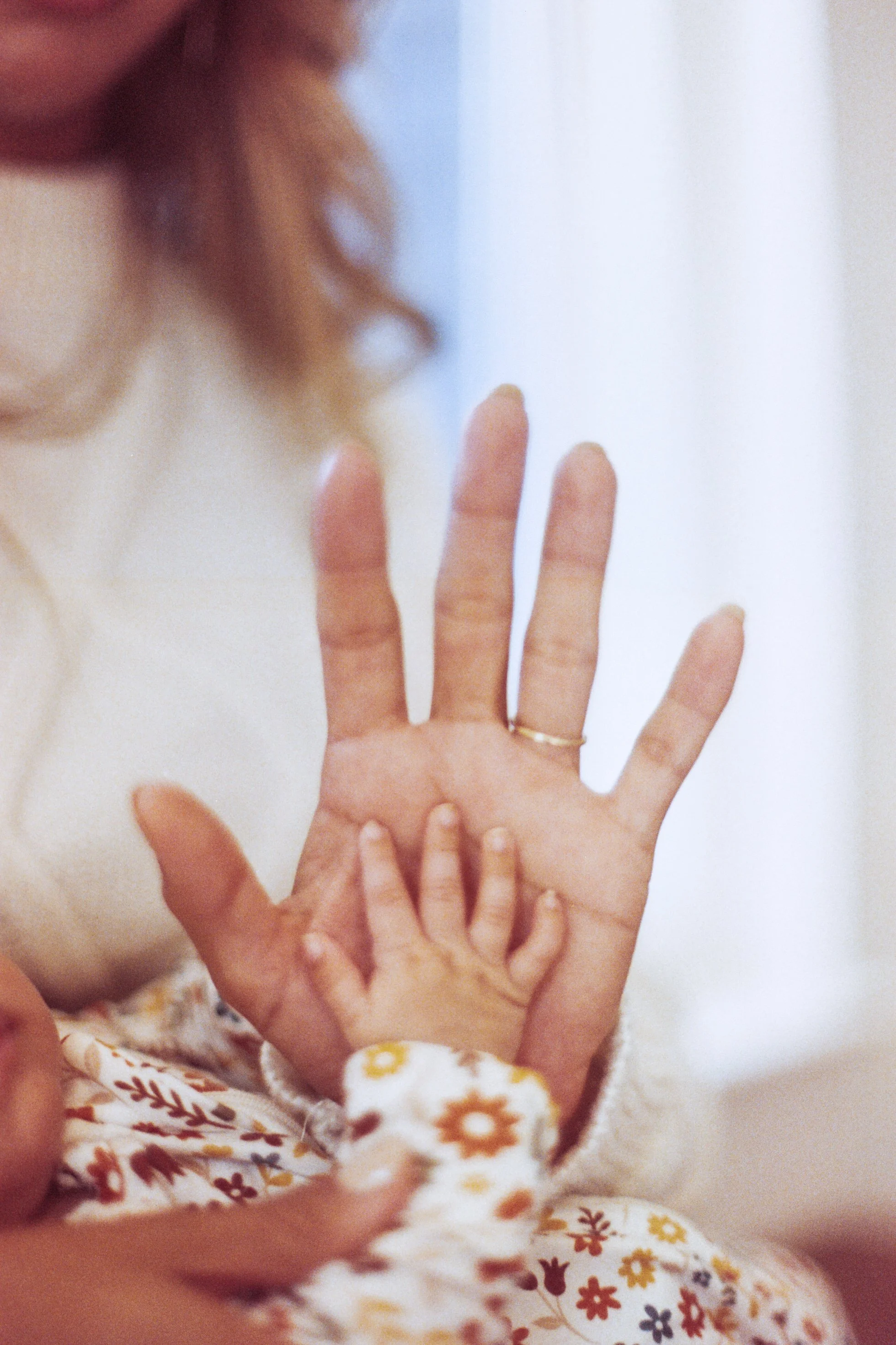 A close-up photo of an adult's hand with a gold ring on the ring finger, placed on a child's head and hand. The child's head is covered with hair, and a smaller child's hand is touching the adult's hand. The background shows a person's face, possibly