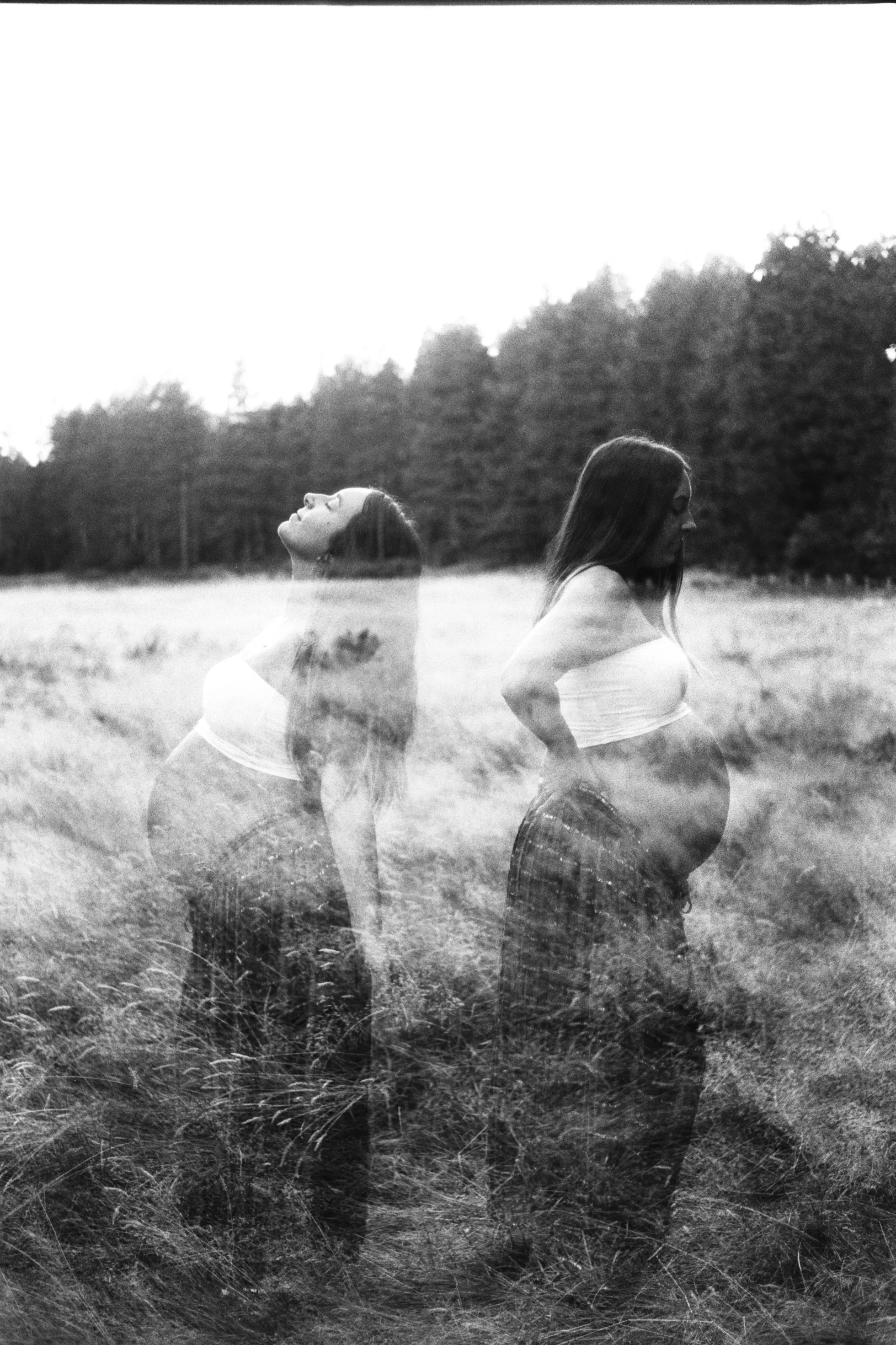 Black and white photo of two women in a field, one with eyes closed and head tilted back, and the other with head down and side profile visible, with trees in the background.