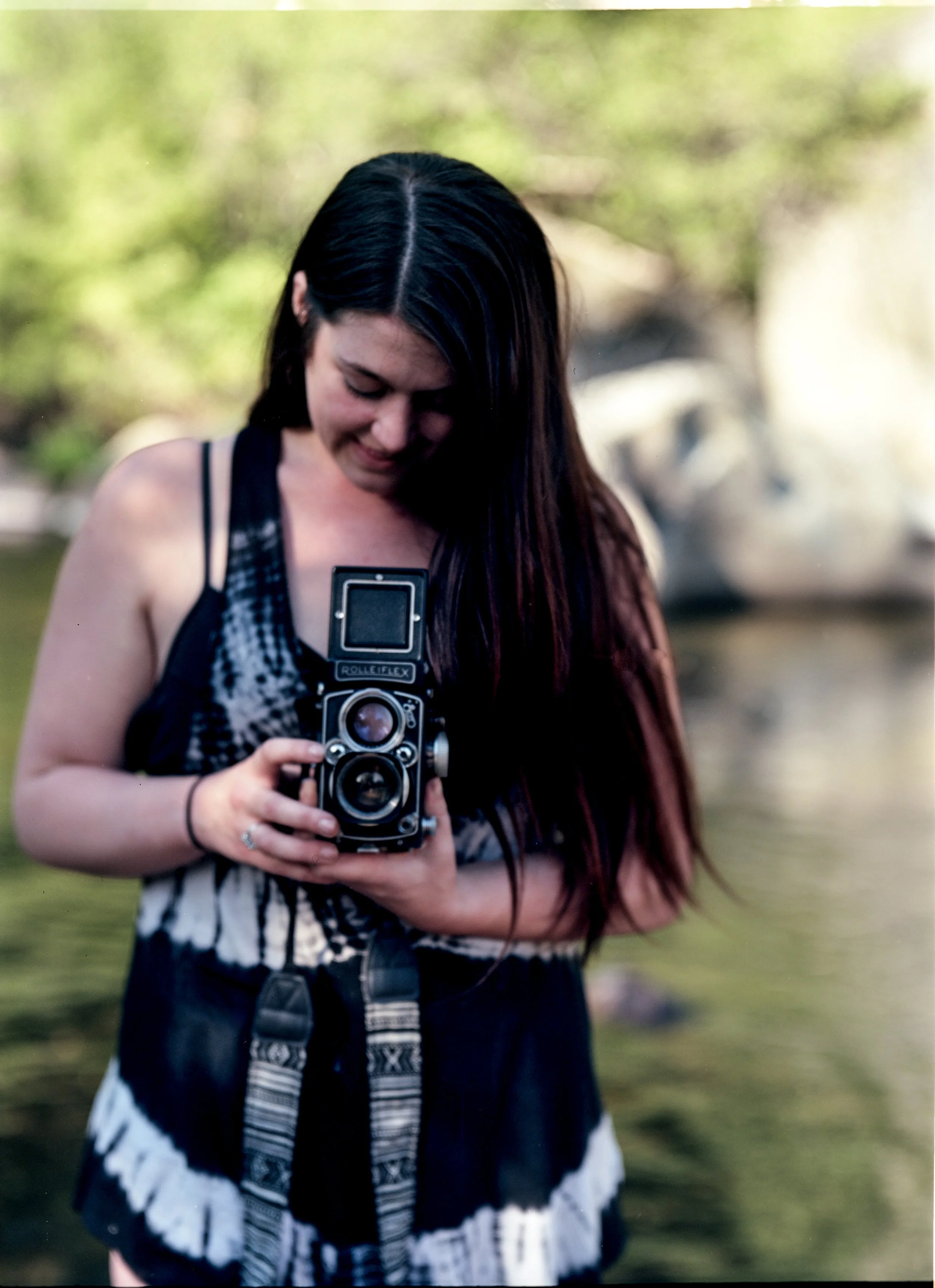 A young woman with long dark hair wearing a sleeveless top and a patterned skirt holding a vintage Rolleiflex camera outdoors in a blurred natural setting with green foliage in the background.