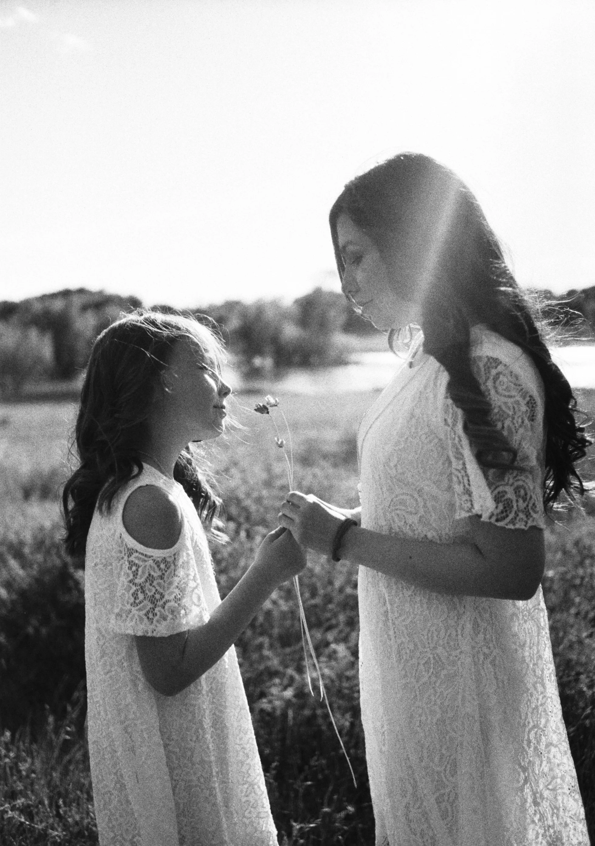 A woman and a young girl are standing outdoors, holding hands, with the woman giving the girl a flower. They are both wearing lace dresses, and the scene is backlit, creating a soft, emotional atmosphere.