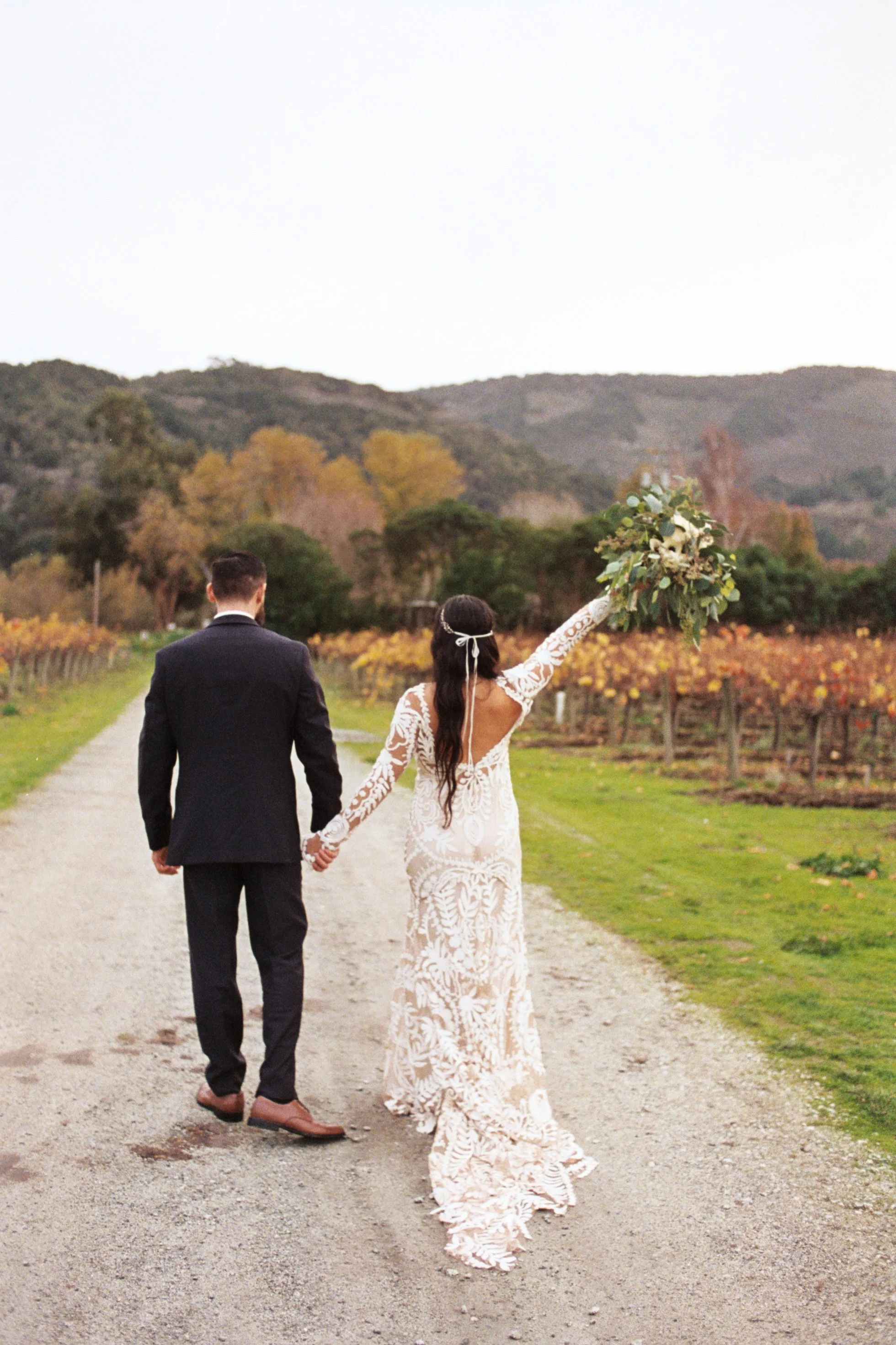 A bride and groom walking hand in hand through a vineyard, with the bride holding a bouquet and wearing a white lace wedding dress, and the groom in a dark suit, during the daytime with hills and trees in the background.