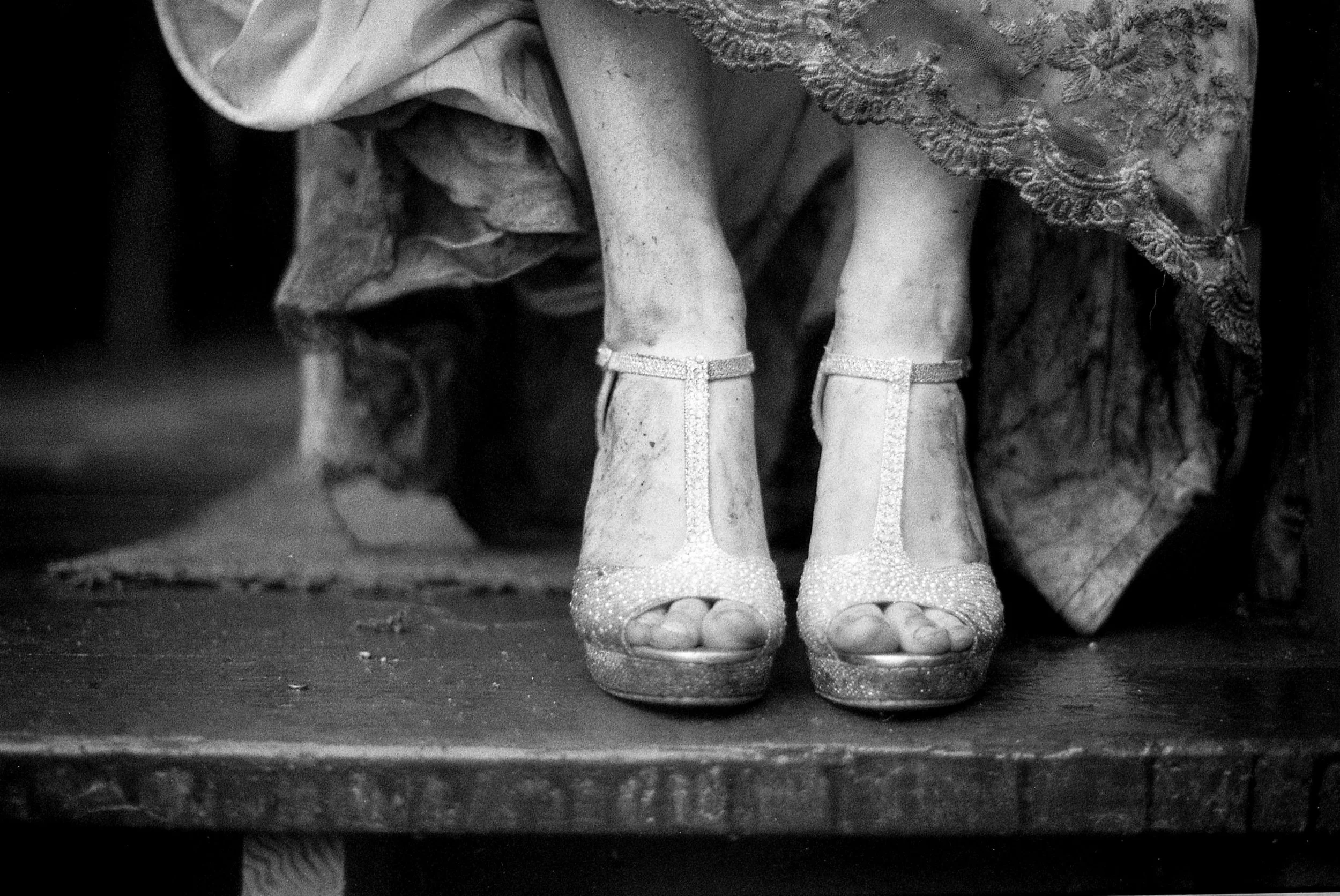 Close-up of a person's feet wearing high-heeled sandals with ankle straps, standing on a reflective surface with another person's feet partially visible in the background.