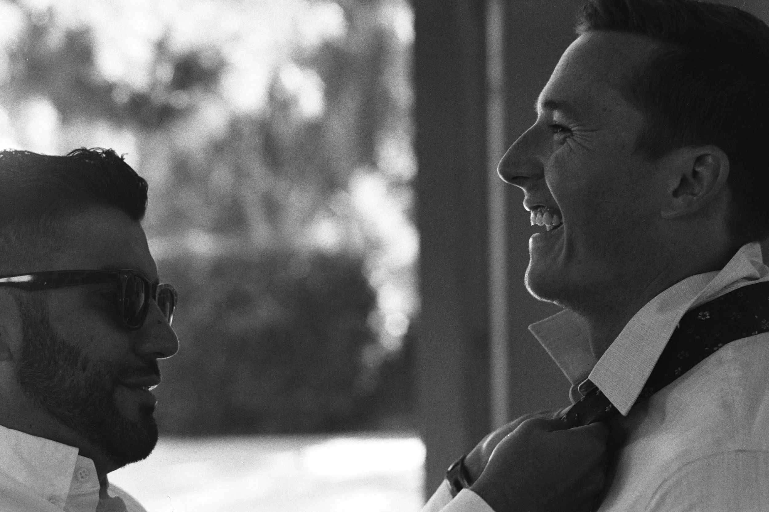 A black-and-white photo of two men smiling and facing each other closely, with one man on the left wearing glasses and a collared shirt, and the other man on the right with a short haircut, dressed in a shirt and blazer, in an outdoor setting.