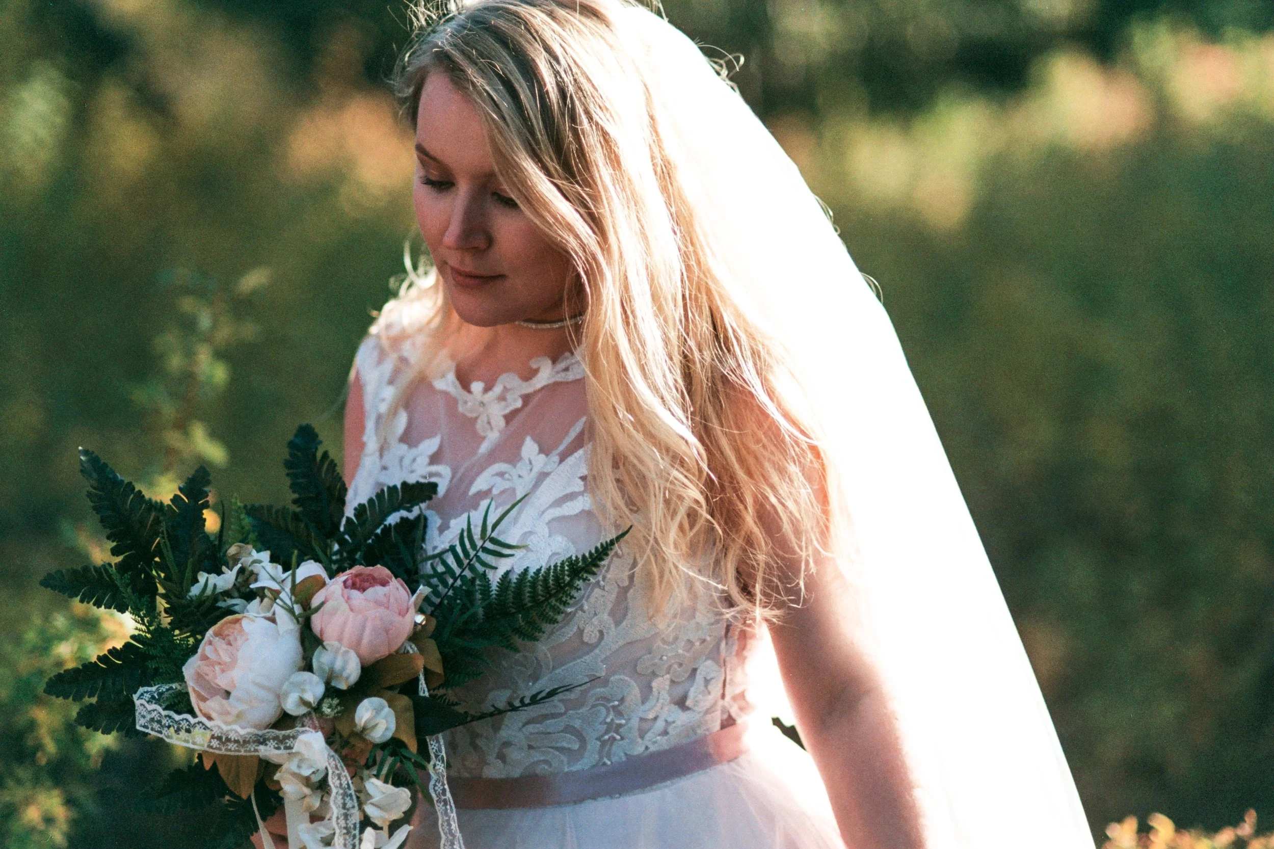 A woman in a white lace wedding dress holding a bouquet of pink and white flowers with greenery, outdoors with sunlight and blurred green trees in the background.