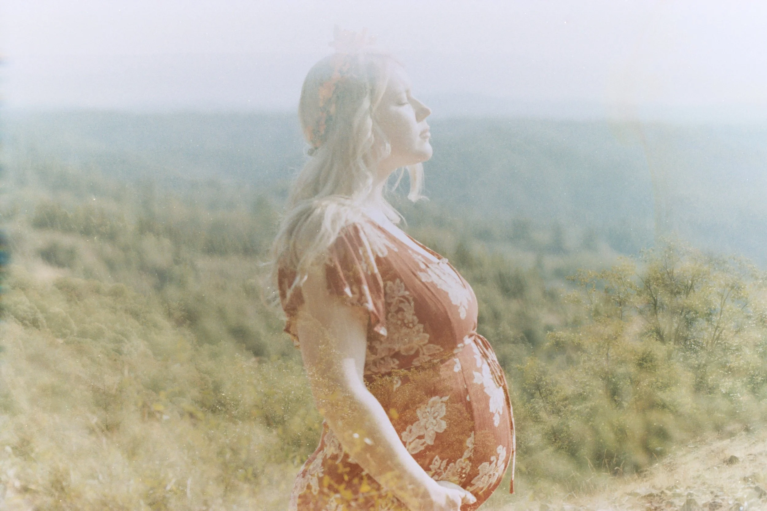 A woman with closed eyes standing outdoors in a field of grass and trees, wearing a floral red dress.