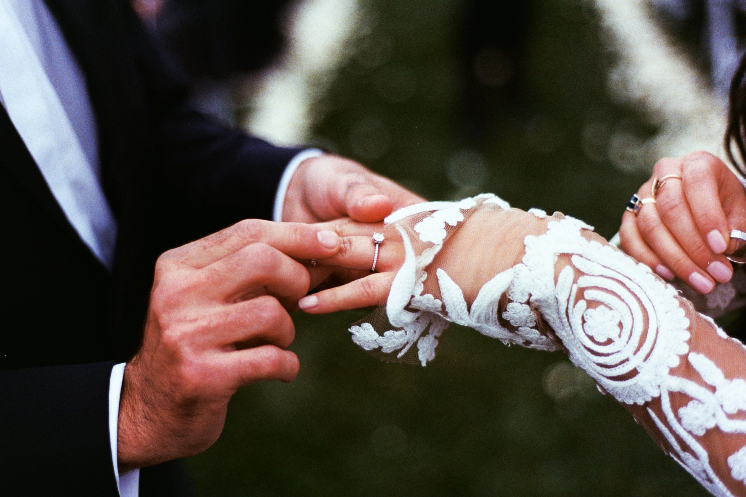 A close-up of a wedding ceremony showing a groom placing a ring on the bride's finger, with the bride wearing a lace dress and rings on her fingers.