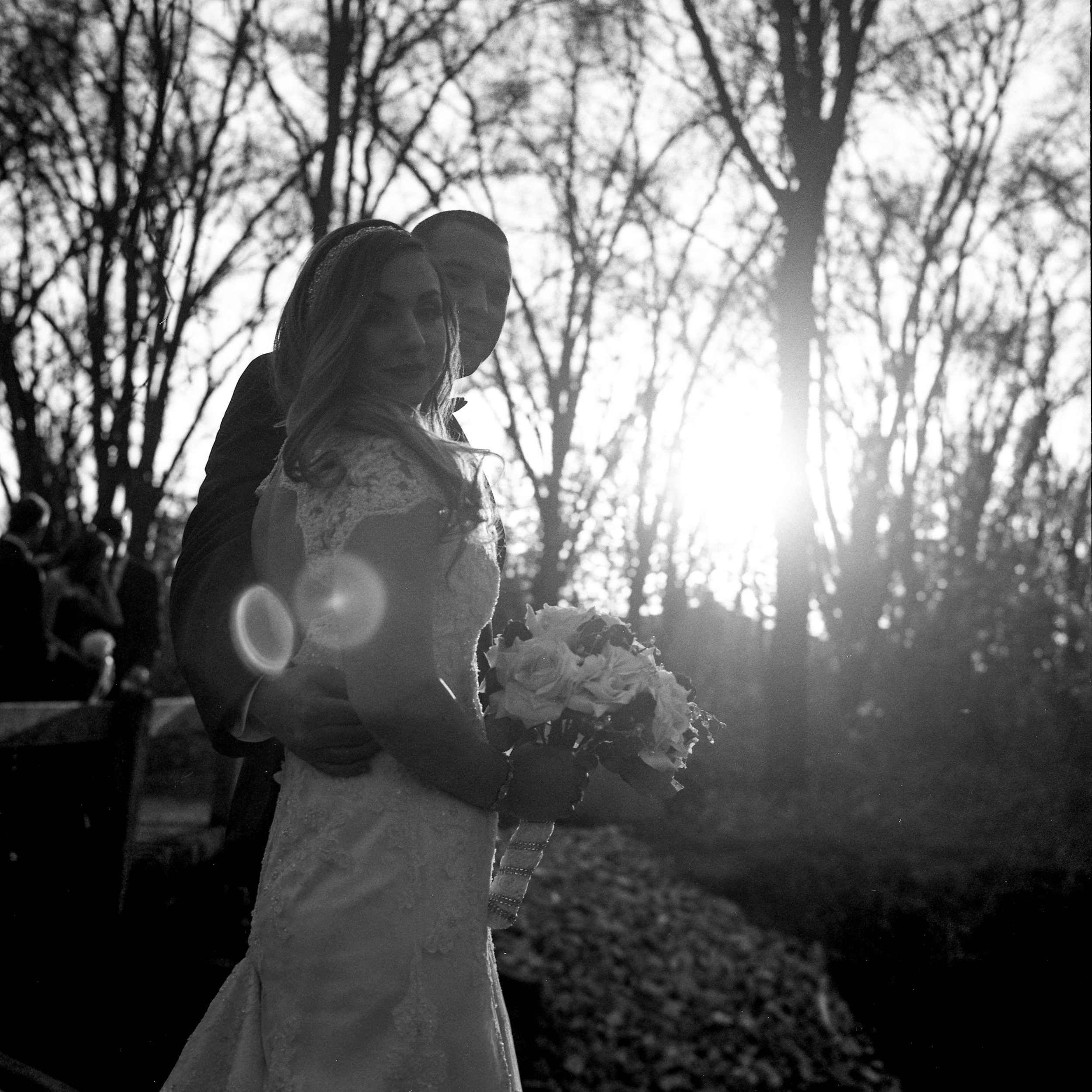 Child holding a bouquet of flowers outdoors in black and white, with bare trees in the background and sunlight shining through.