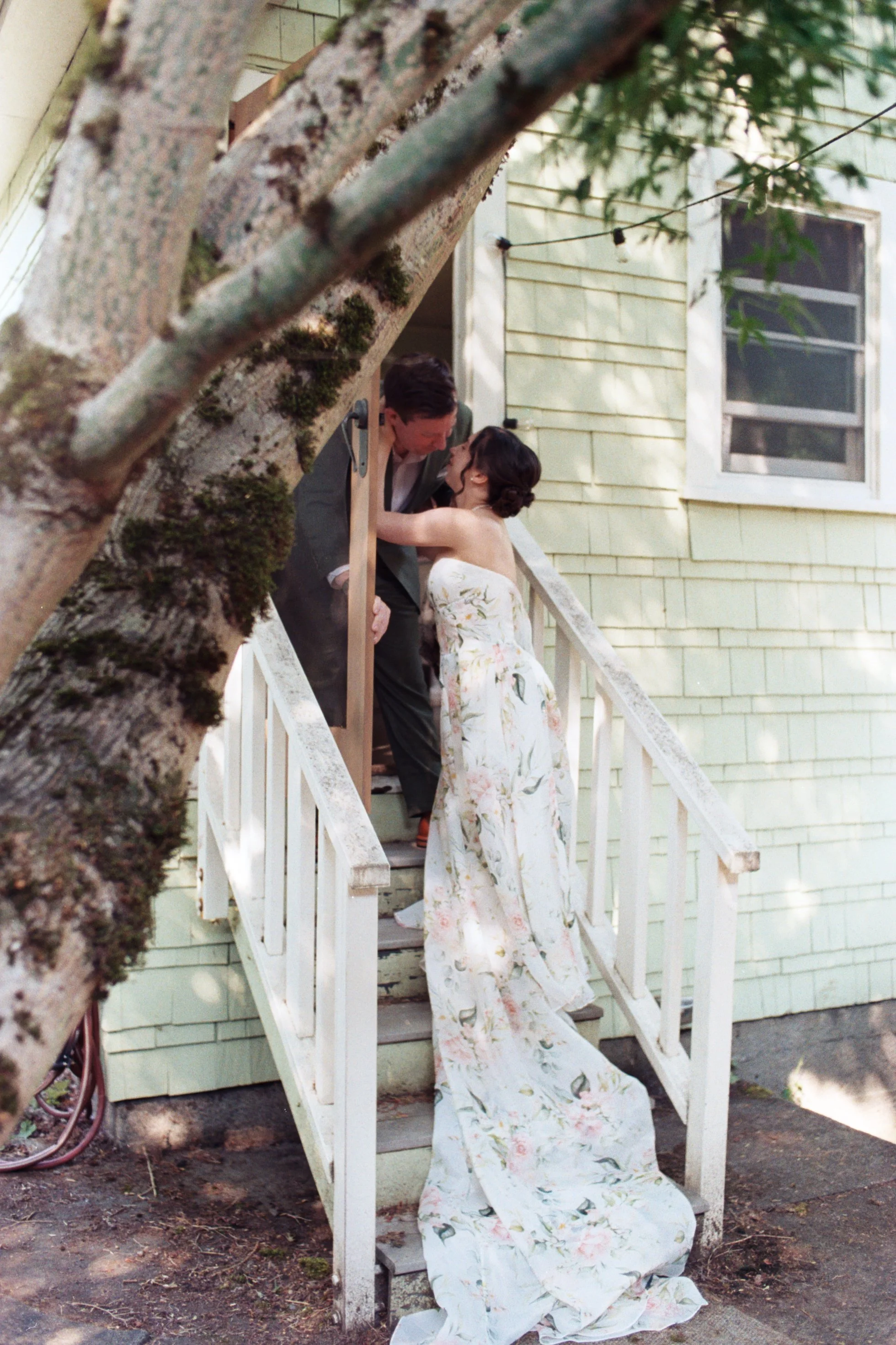 A woman in a floral dress is kneeling at the bottom of a staircase, while a man in a gray blazer and white shirt is standing on the stairs, leaning down to her, both smiling and engaging each other. The background shows a yellow house with a window, 
