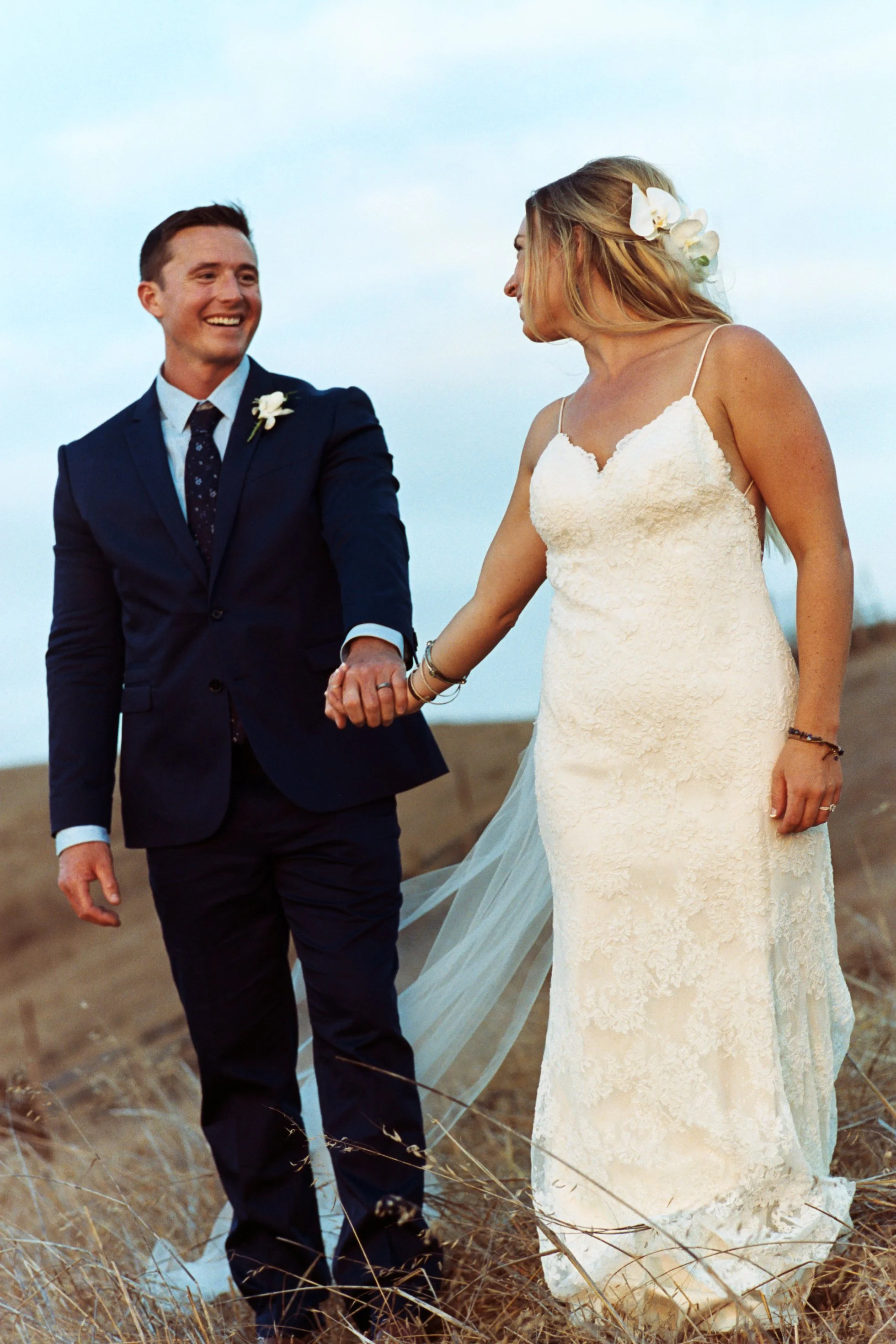 A happy couple holding hands in a field during an outdoor wedding, with the woman in a white wedding dress and the man in a dark suit.