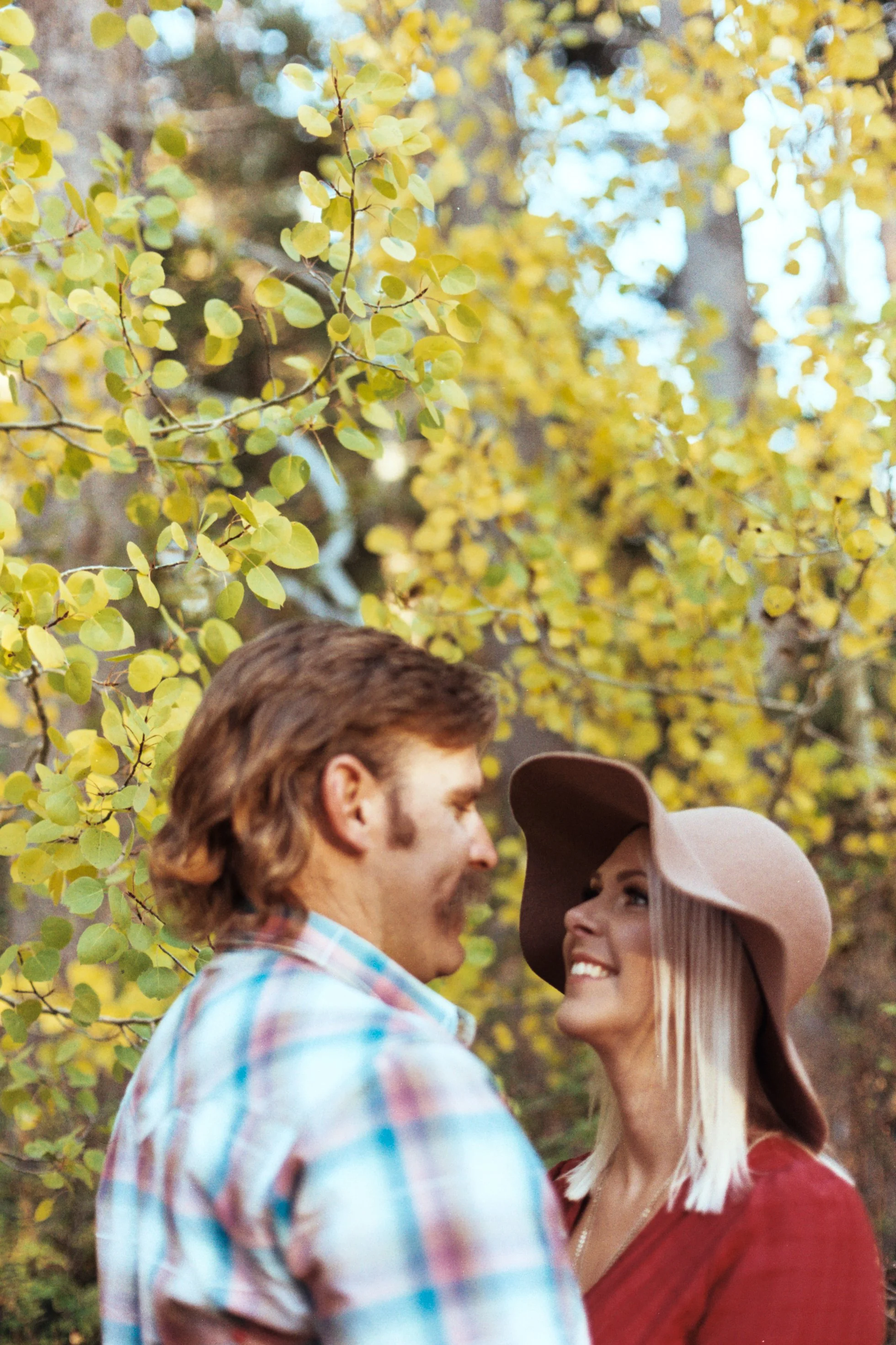A man and woman face each other smiling outdoors with yellow autumn leaves in the background.
