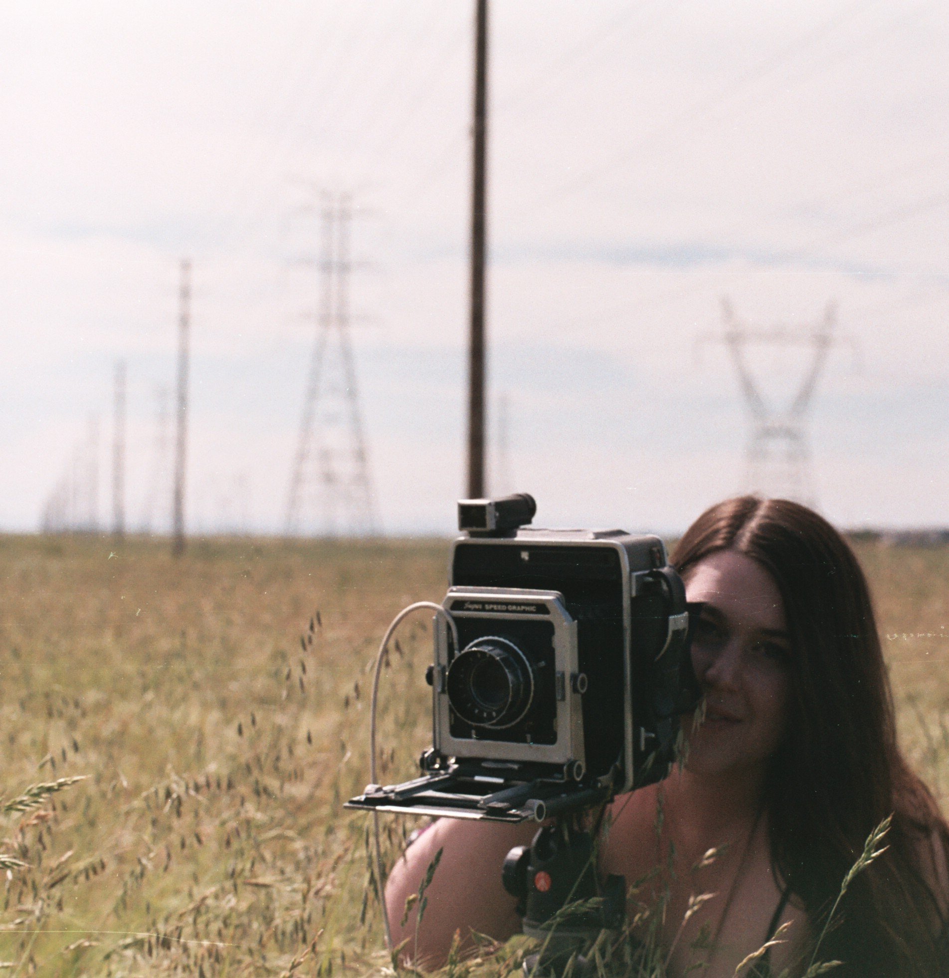 A person lying on the ground in an open field, taking a photo through a camera mounted on a stabilizing rig, with power line towers in the background and a cloudy sky.
