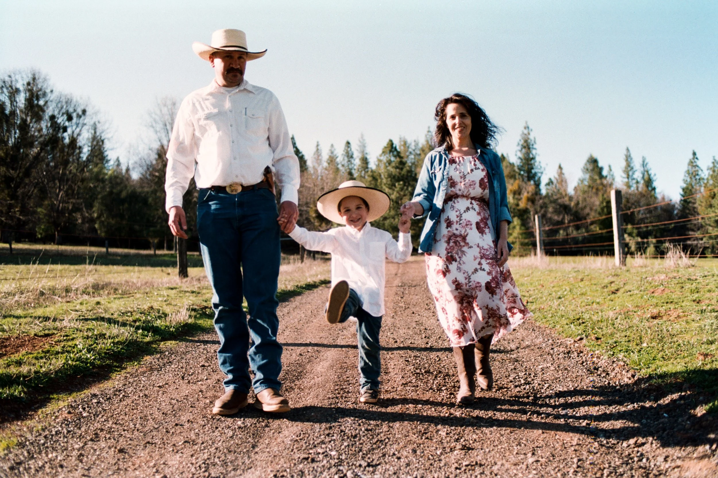 A family of three walking outdoors on a dirt path in a rural setting, with the father wearing a cowboy hat and white shirt, the mother in a floral dress and denim jacket, and the child in a large straw hat and white shirt, smiling and holding hands.