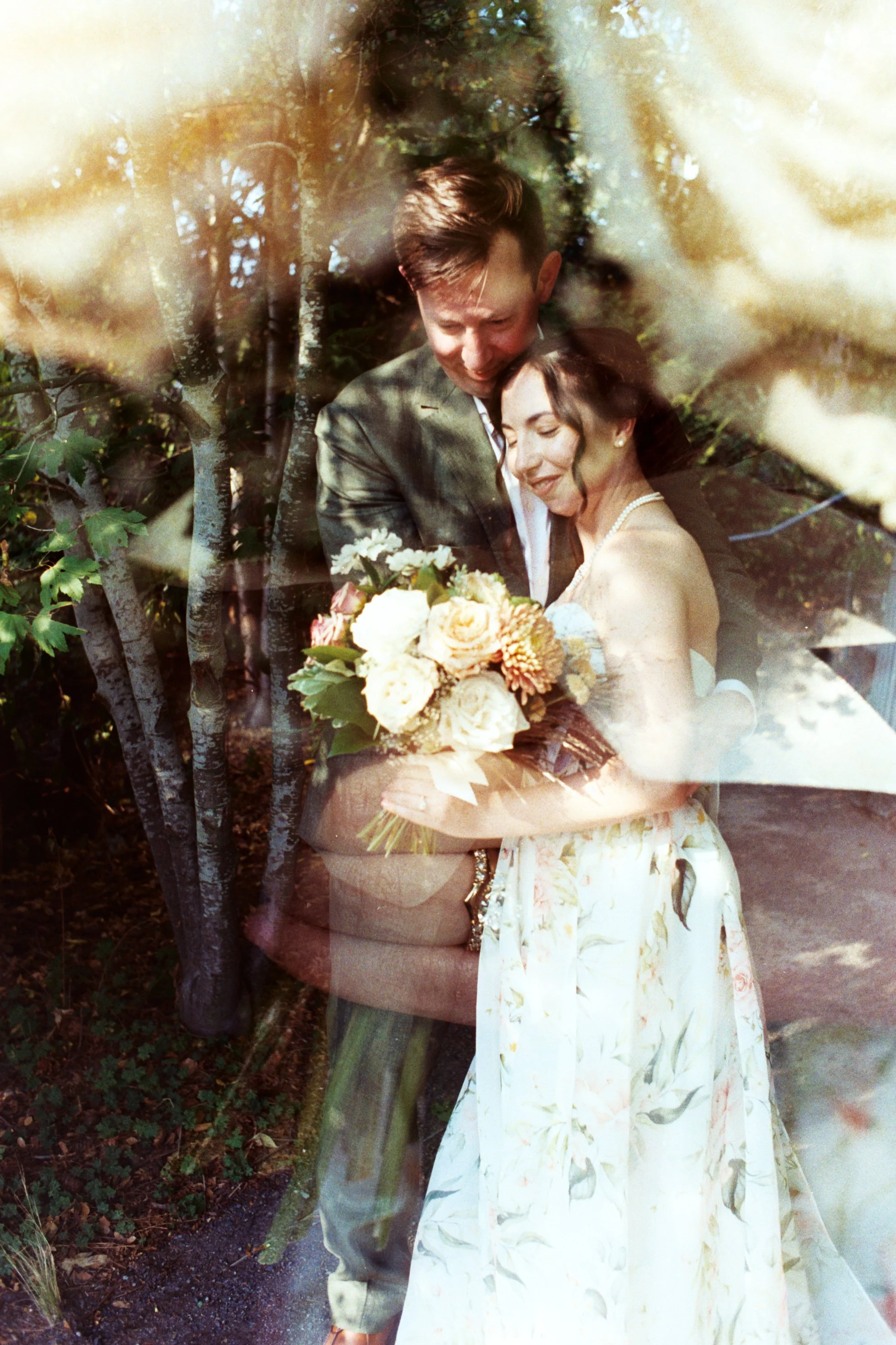 A couple dressed in wedding attire sharing a tender moment outdoors, surrounded by trees, with the woman holding a bouquet of flowers.