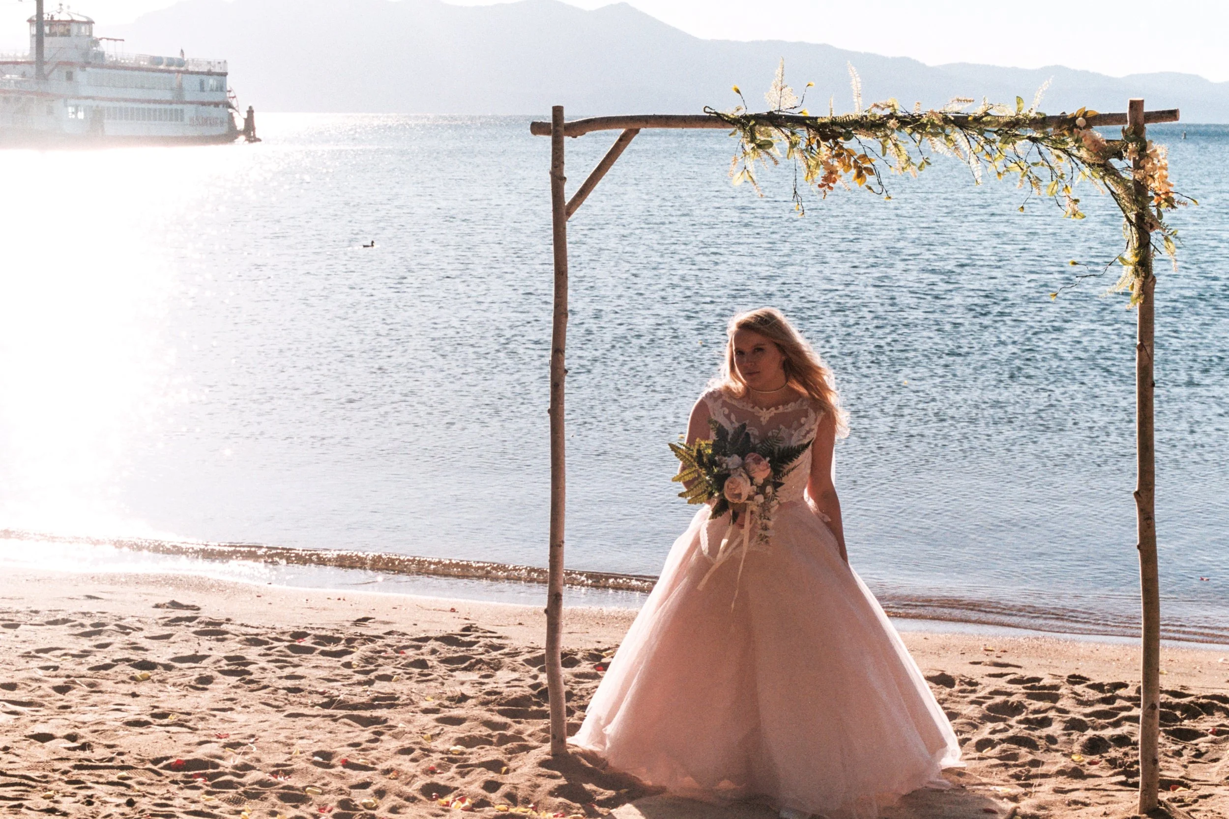 A woman in a wedding dress holding a bouquet sitting on a sandy beach beneath a wooden arch decorated with flowers and greenery, with water and mountains in the background.