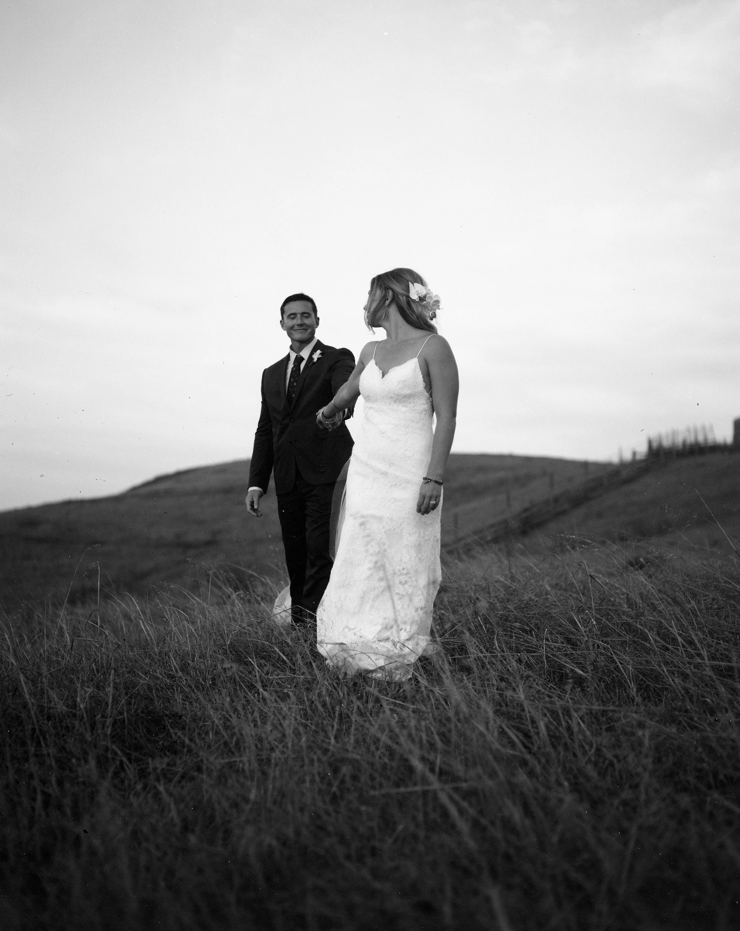 Black and white photo of a bachelorette couple walking through grassy fields on a hill, one woman in a white wedding dress and flower crown, and a man in a suit, smiling and holding hands.