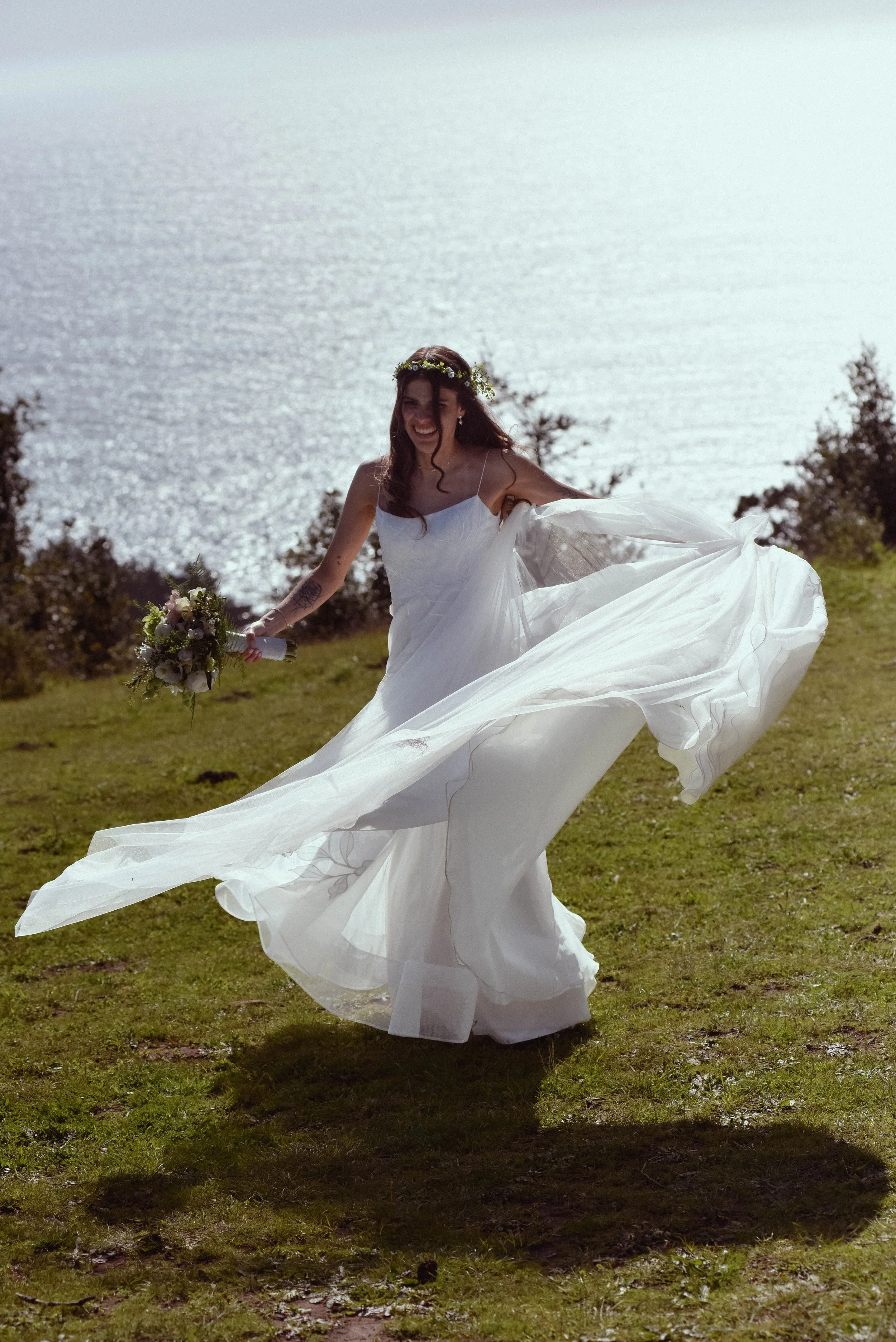 A woman in a white wedding dress holding a bouquet of flowers, smiling and playing outdoors on a grassy hill near water.