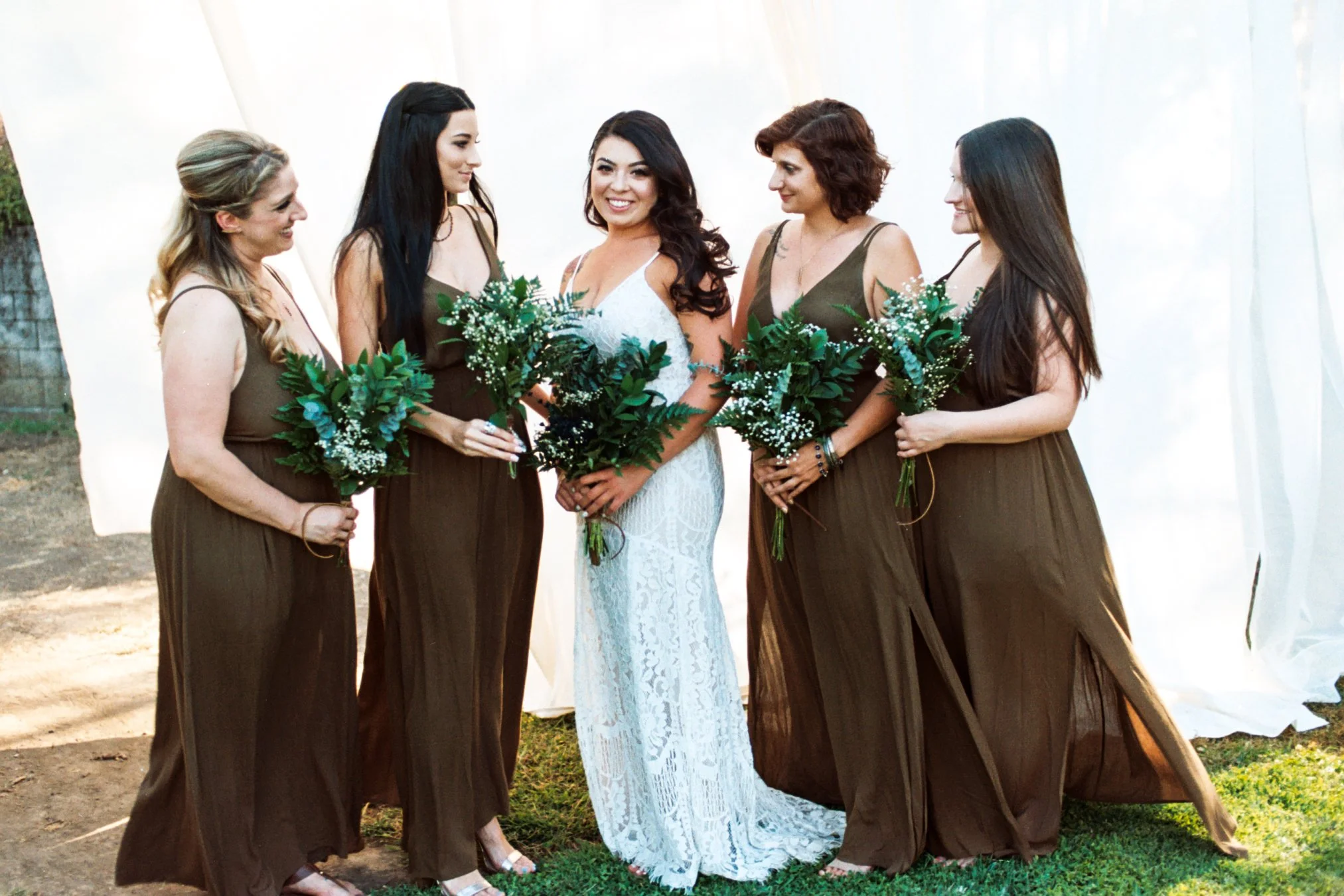 A bride in a white lace dress smiling with four bridesmaids in brown dresses holding bouquets of greenery, standing outdoors in front of a white backdrop.