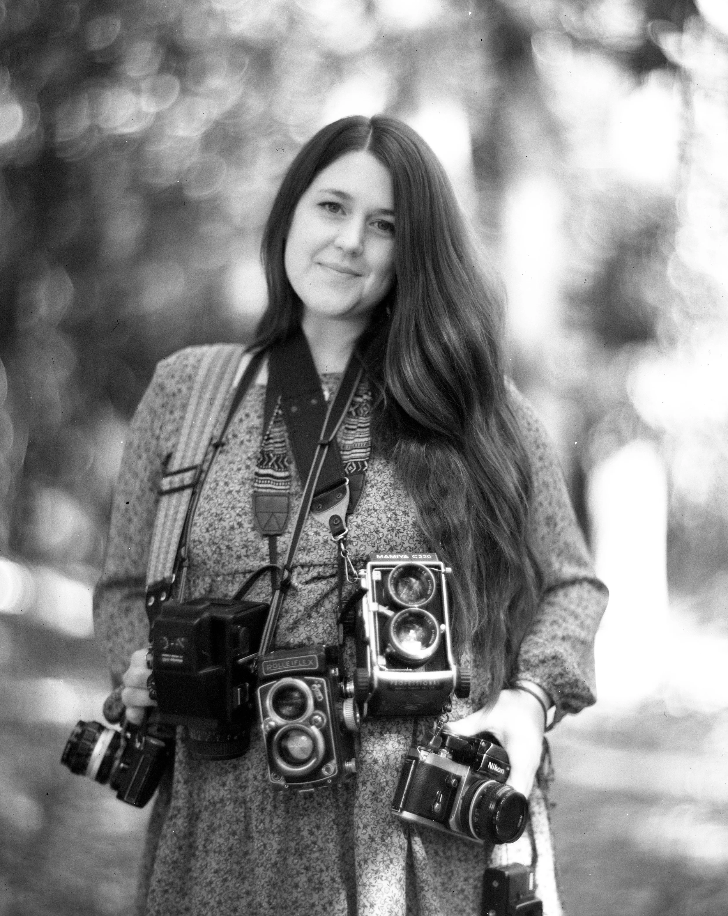 A woman with long dark hair and light skin, wearing a patterned top, is standing outdoors surrounded by blurred bokeh lights. She has multiple cameras hanging from her neck and shoulders, and she is holding a camera in her right hand. She is smiling 