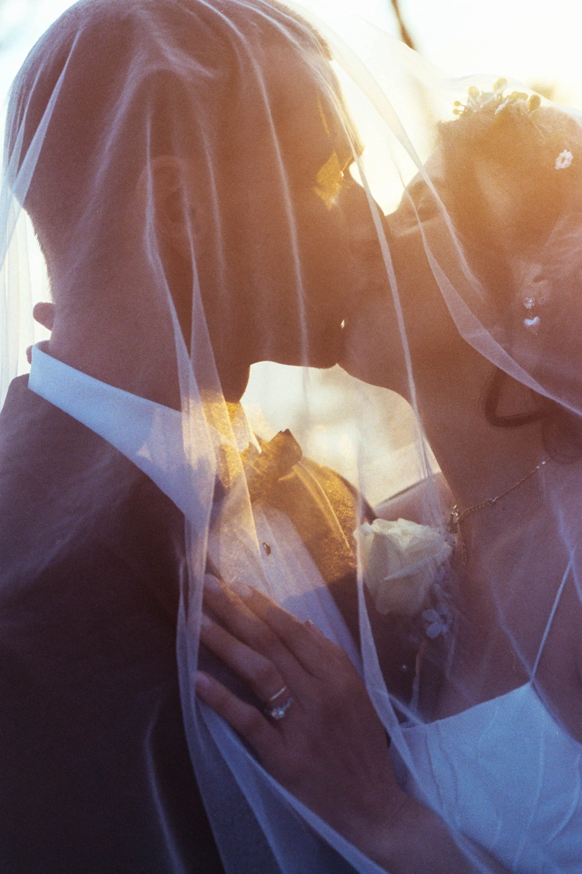A bride wearing a white veil and pearl earrings, kissing her groom who is dressed in a dark tuxedo with a white shirt and bow tie, during their wedding ceremony.