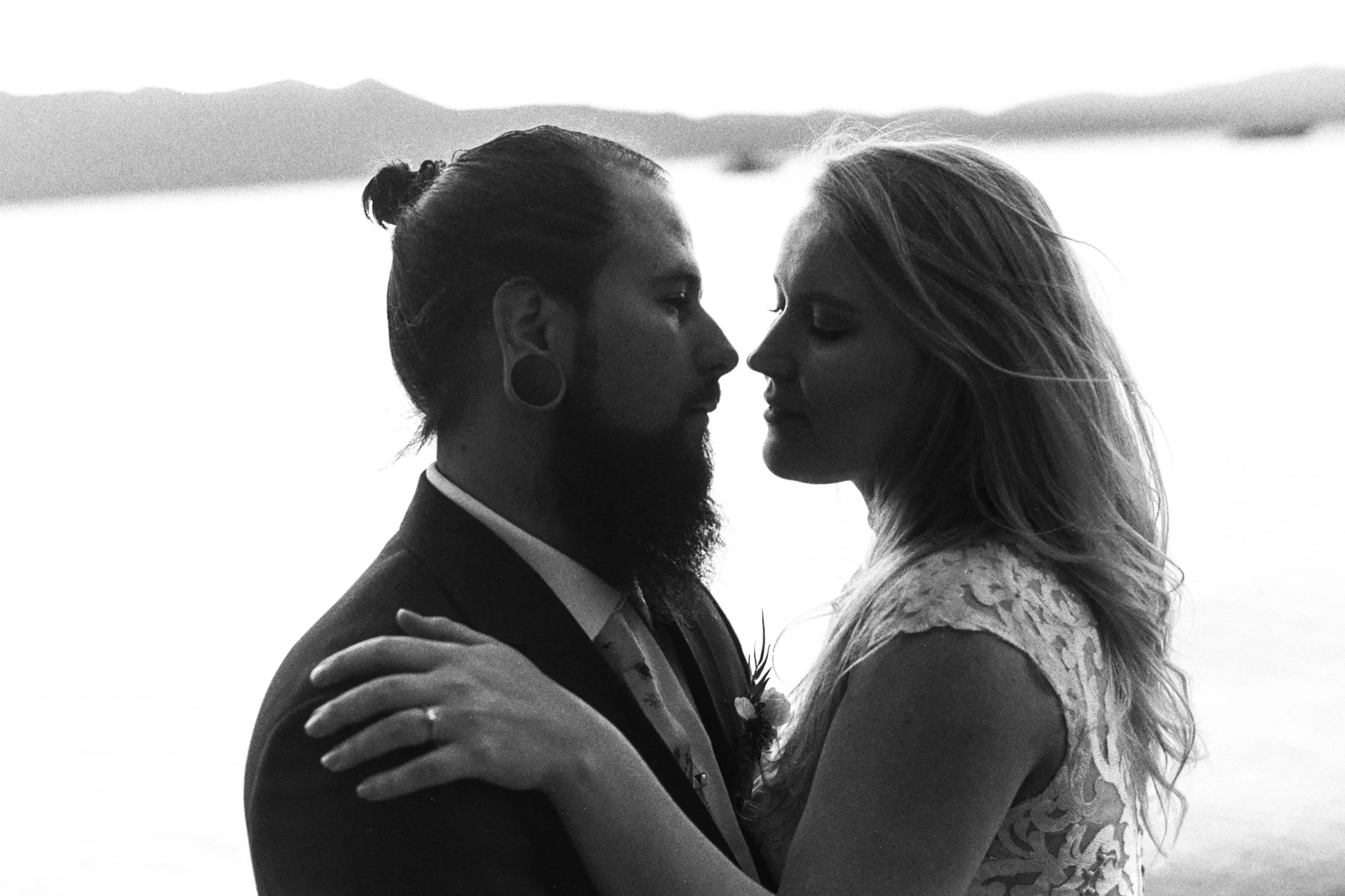 Black and white photo of a couple embracing each other closely with their foreheads touching, outdoors with a blurred landscape in the background.