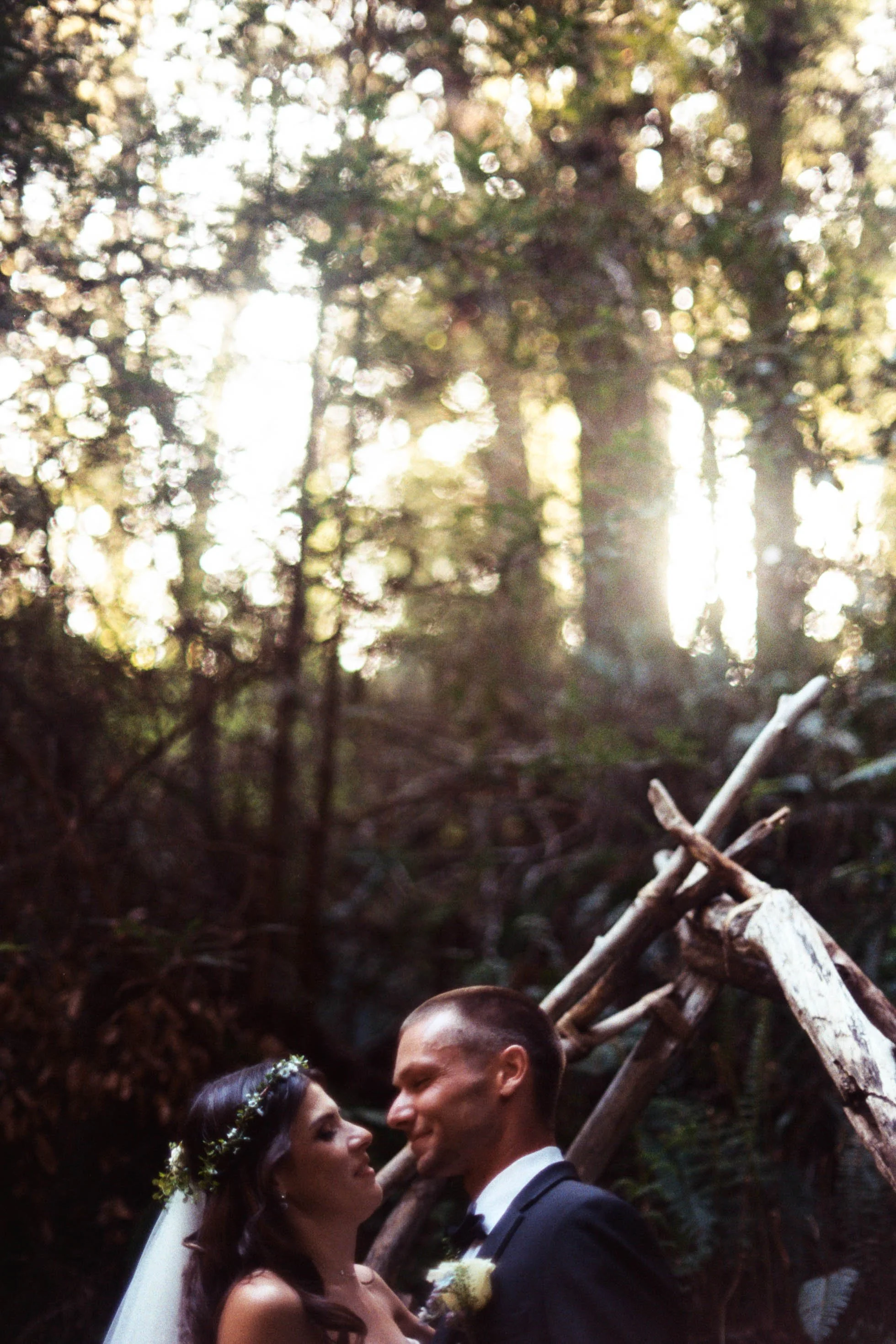 A bride and groom, dressed in wedding attire, face each other with noses nearly touching during an outdoor wedding ceremony in a wooded area. The bride wears a floral headpiece and has long, dark hair, while the groom has short, dark hair and is wear