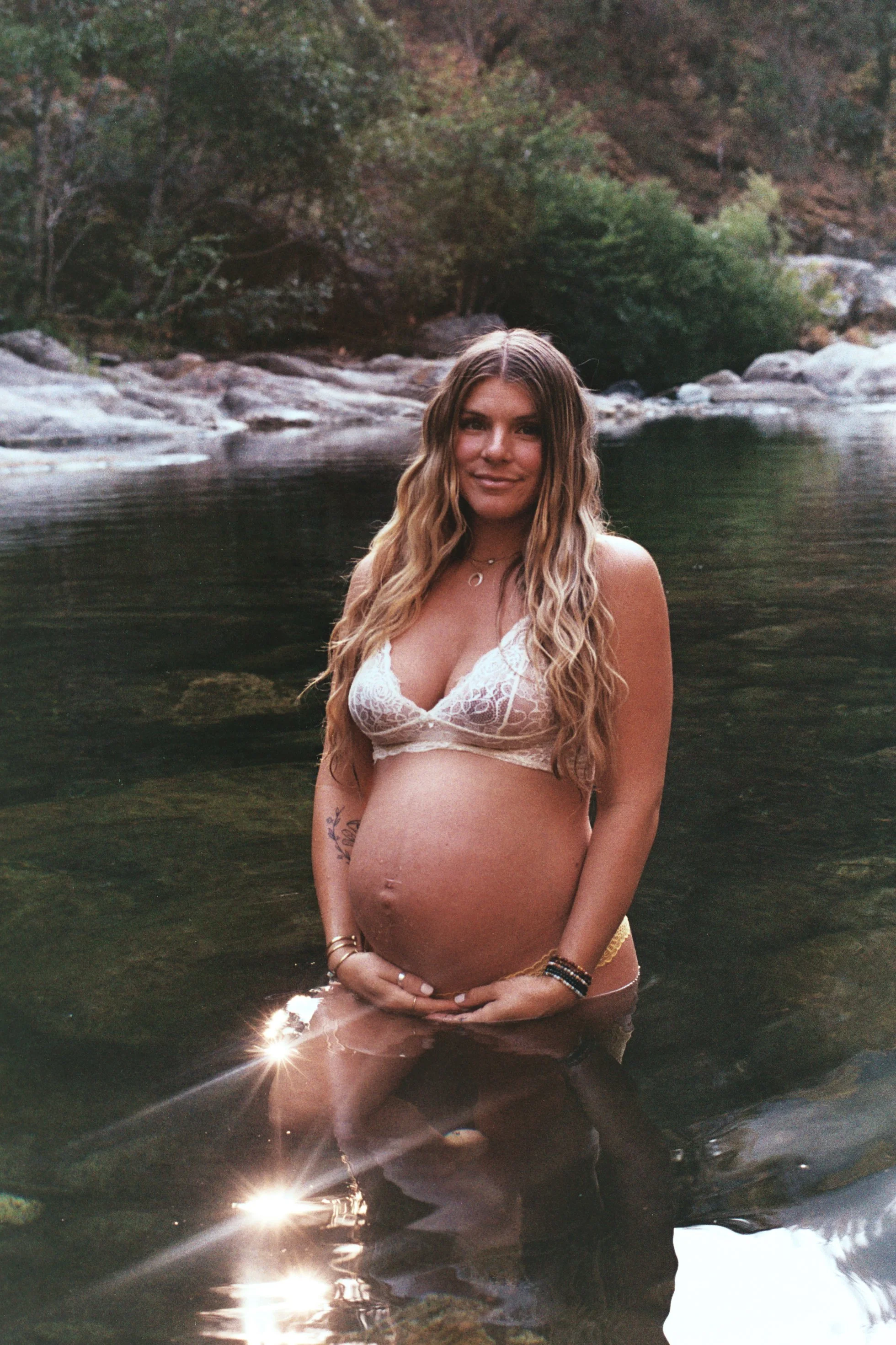 Pregnant woman standing in a river, surrounded by rocks and trees, with the reflection of sunlight on the water.