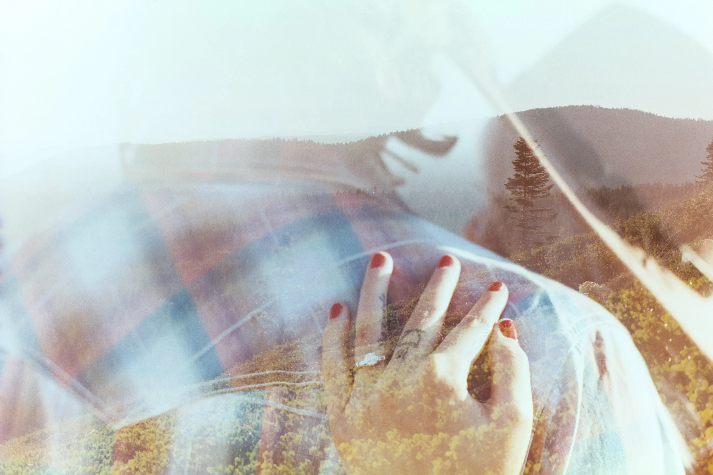 A person's hand with red-painted nails touching a surface covered with yellow and green leaf-shaped objects, viewed through a transparent, slightly curved surface with reflections, in an outdoor setting with trees and a hilly landscape in the backgro