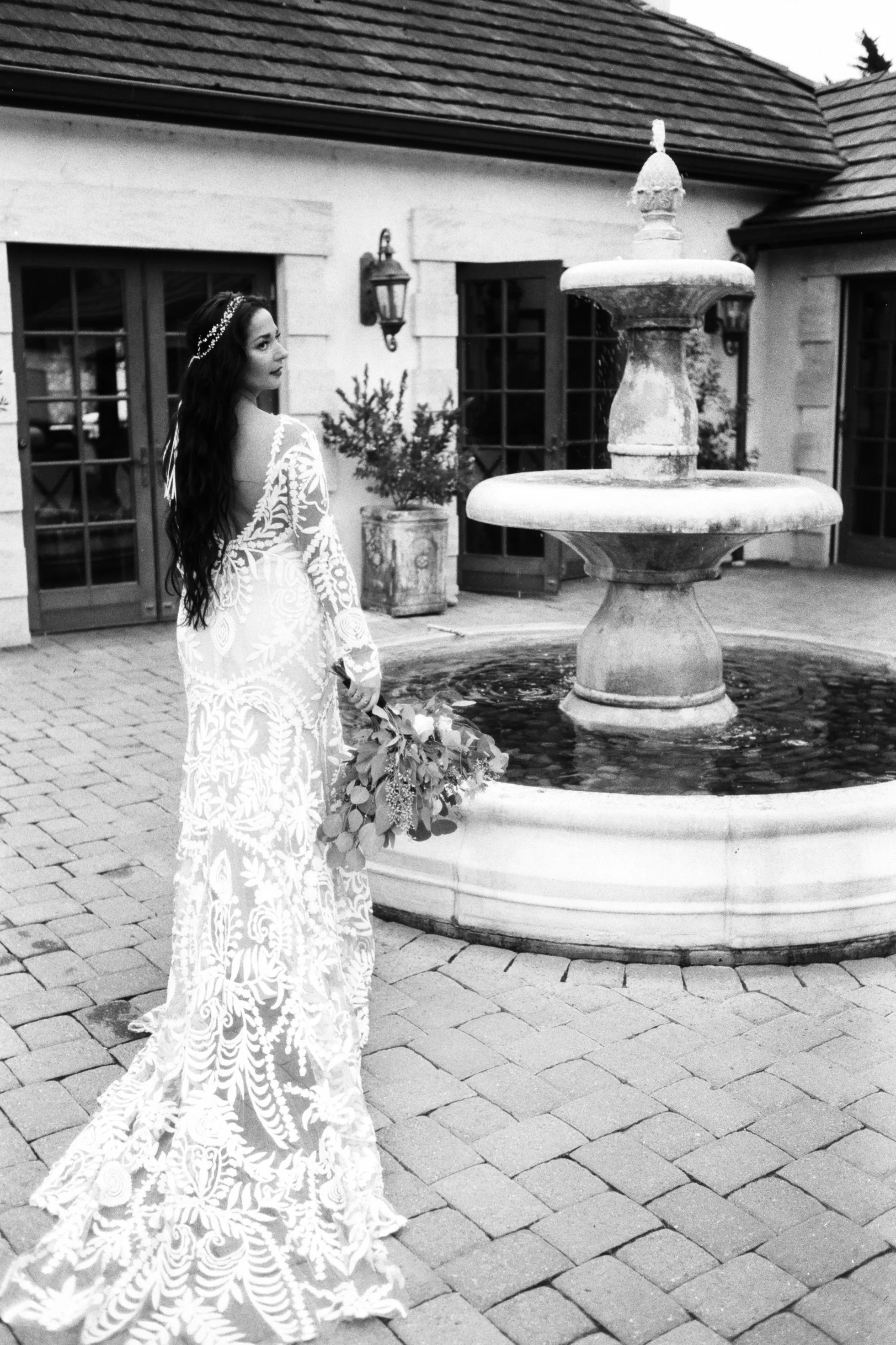 A woman in a white, embroidered dress with long dark hair standing next to a fountain in an outdoor courtyard.