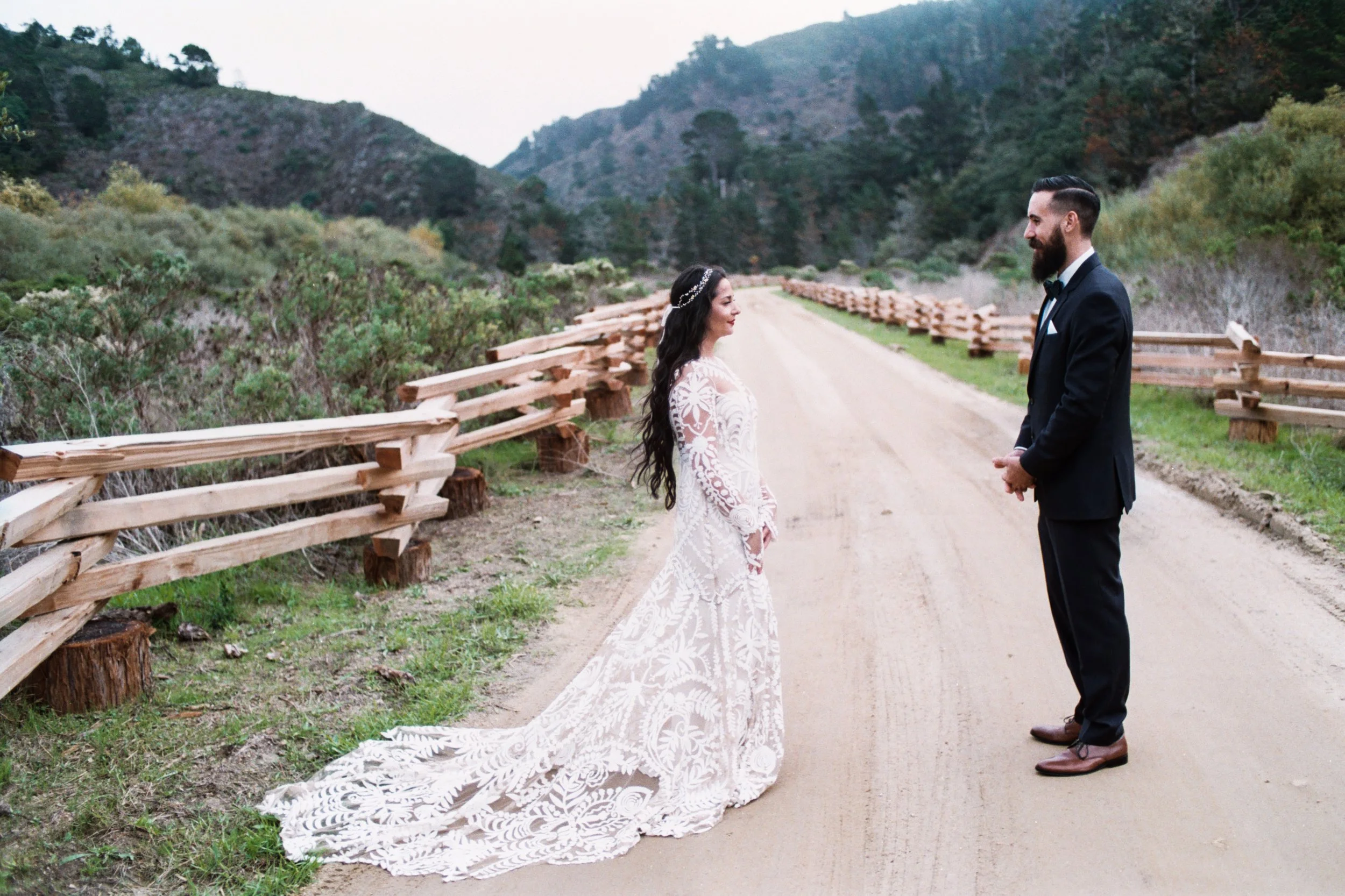 A bride and groom standing on a dirt road with a wooden fence and mountainous landscape in the background. The bride wears a white lace wedding gown with a train, and the groom is dressed in a black tuxedo.
