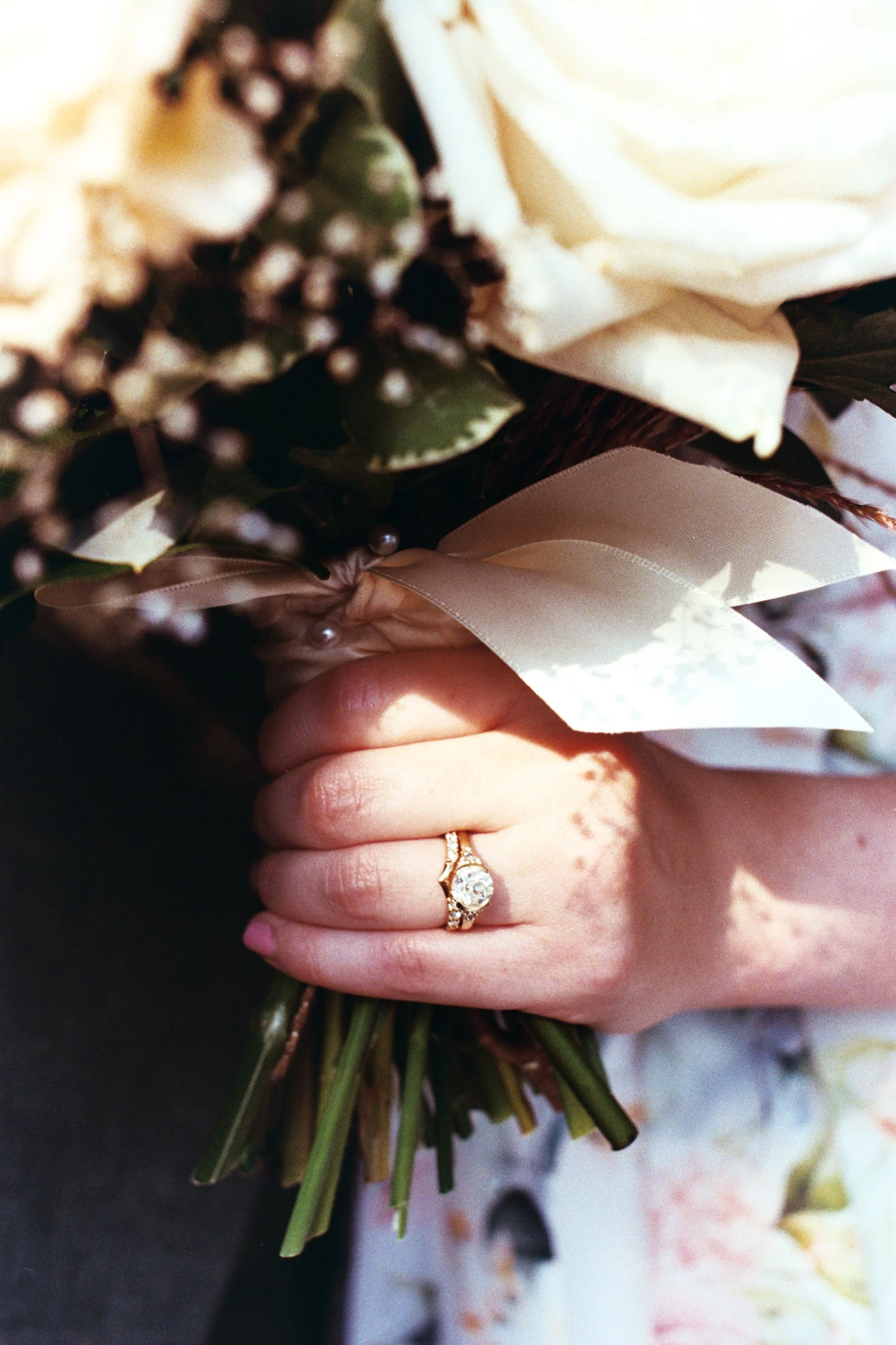 Close-up of a person's hand holding a small bouquet of white flowers with greenery, wearing a gold ring with a large gemstone.