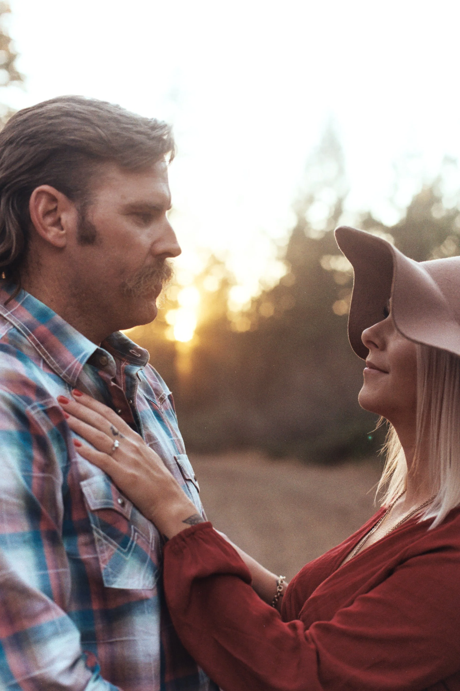A man and woman face each other outdoors during sunset, touching foreheads with eyes closed, sharing a tender moment.