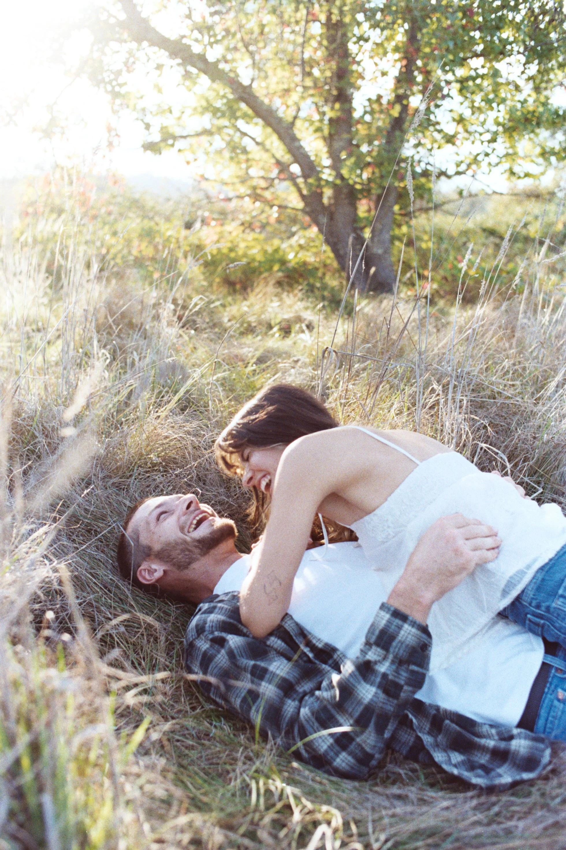 A man and woman lying on grassy ground, smiling, with the woman leaning over the man, outdoors in a field with a large tree and sunlight in the background.