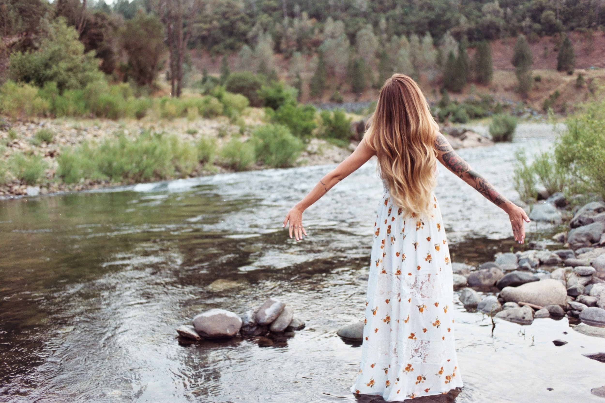 A woman in a white dress with orange floral patterns standing in a shallow river with her arms outstretched, surrounded by greenery and trees in a natural landscape.