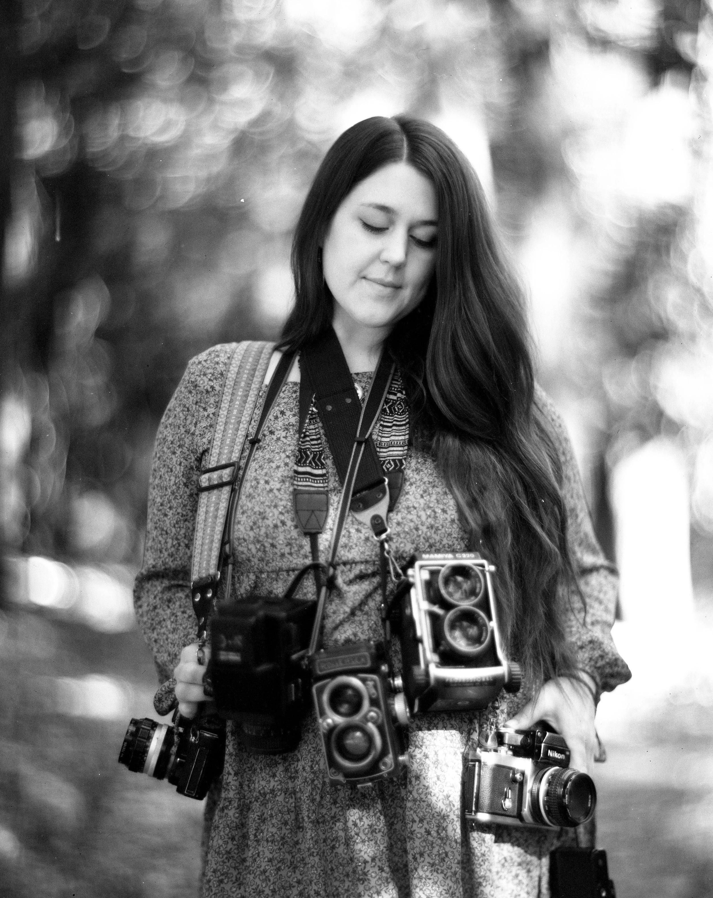 Black-and-white photo of a young woman with long hair, looking down with a gentle smile, holding a vintage camera with multiple lenses, and wearing a patterned dress.
