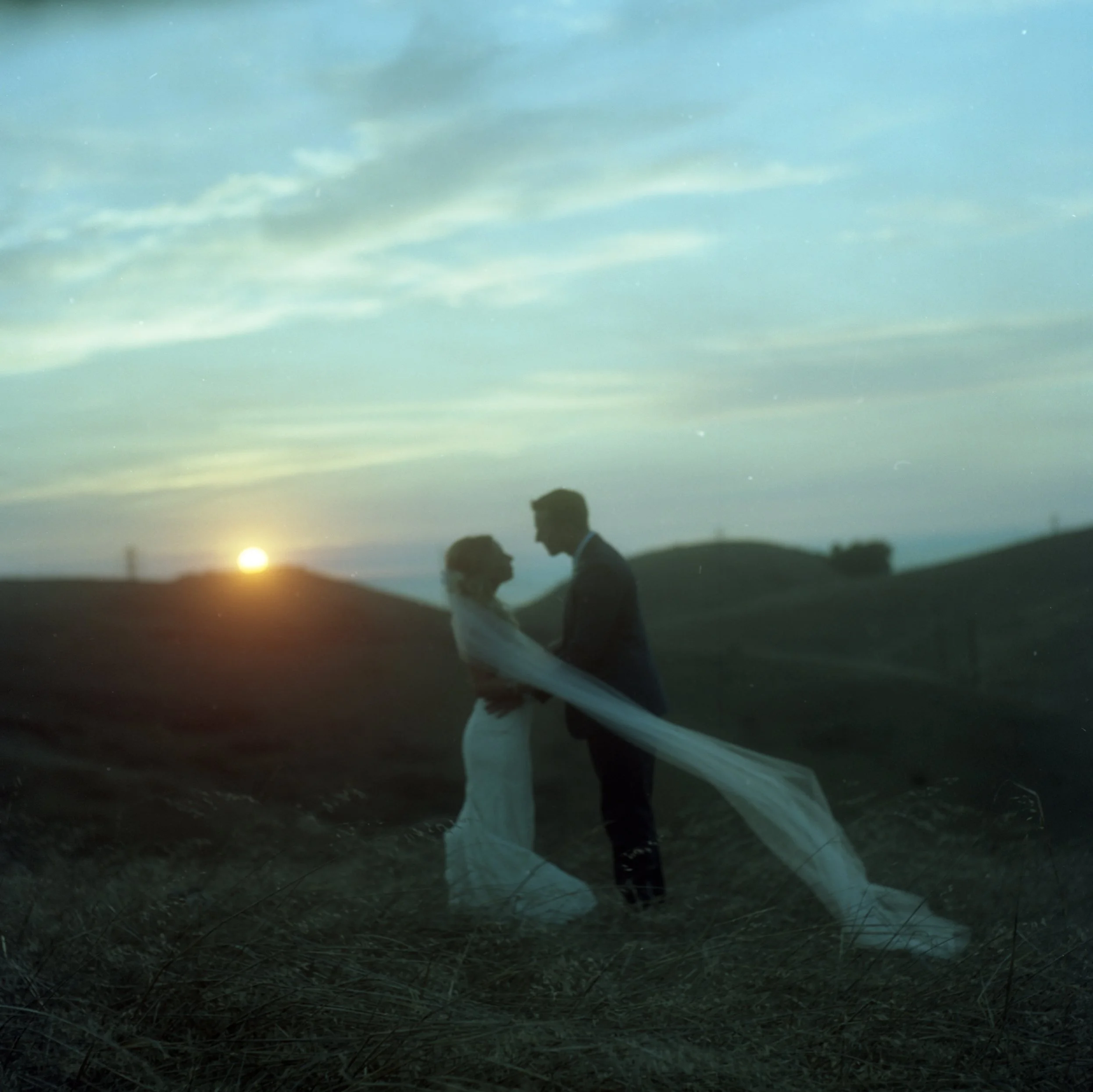 A couple is holding hands and walking on a grassy field during sunset, with the sky showing blues and clouds.