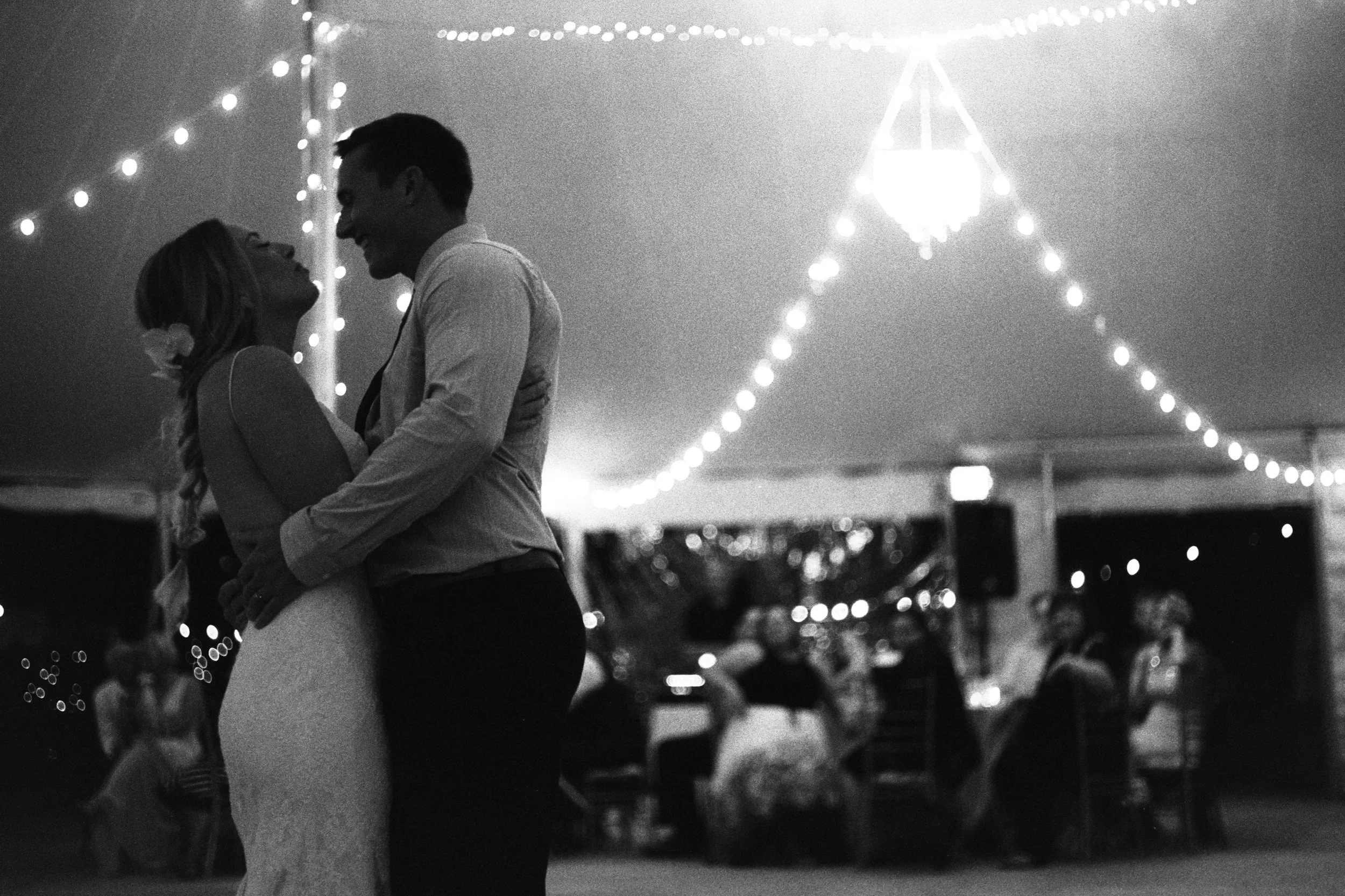 A black and white photo of a couple dancing closely at night under string lights, with a blurred background of tents and people at an outdoor event.
