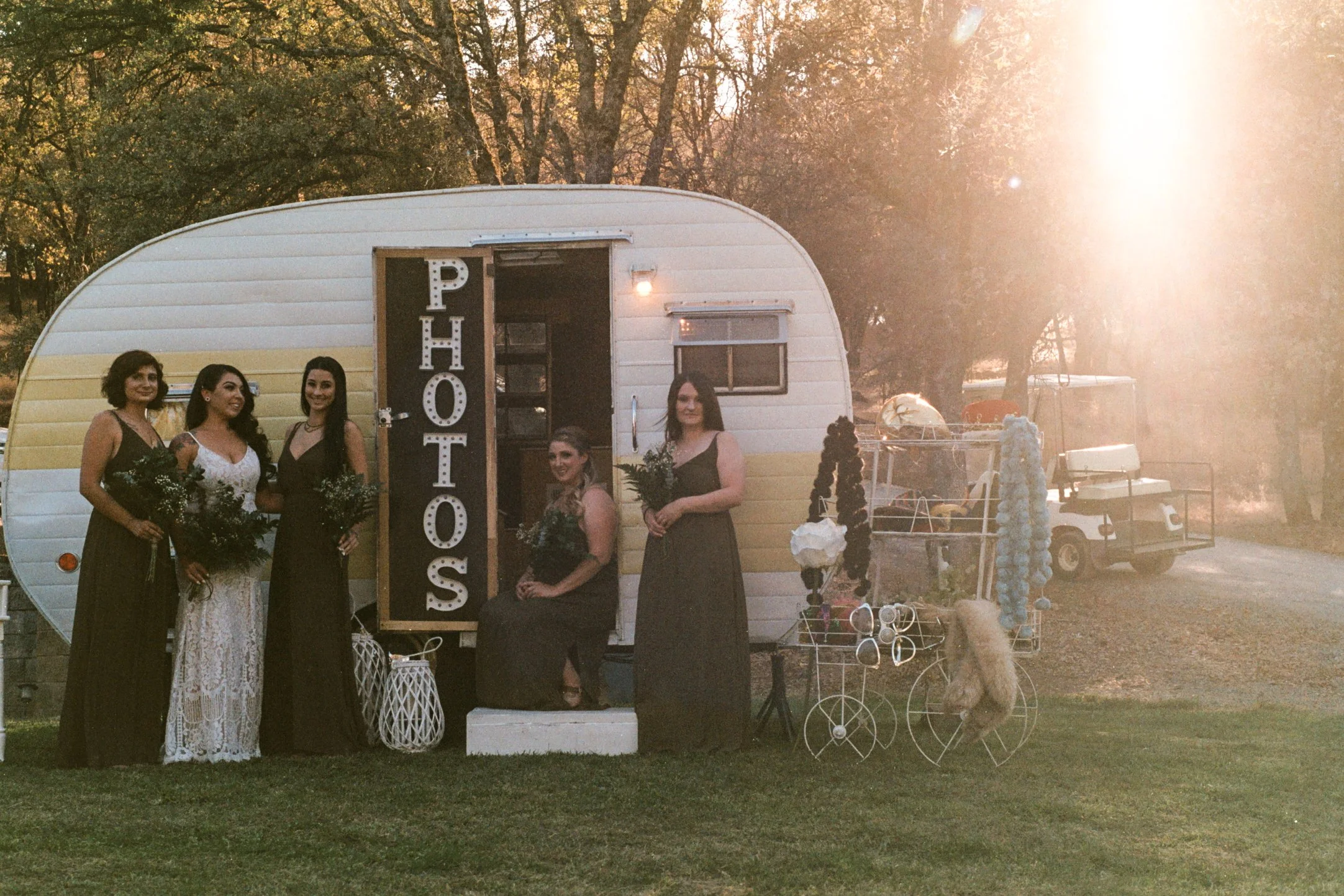 A group of five women in formal dresses standing in front of a vintage trailer with a sign reading 'Photo Booth,' at an outdoor event during sunset.
