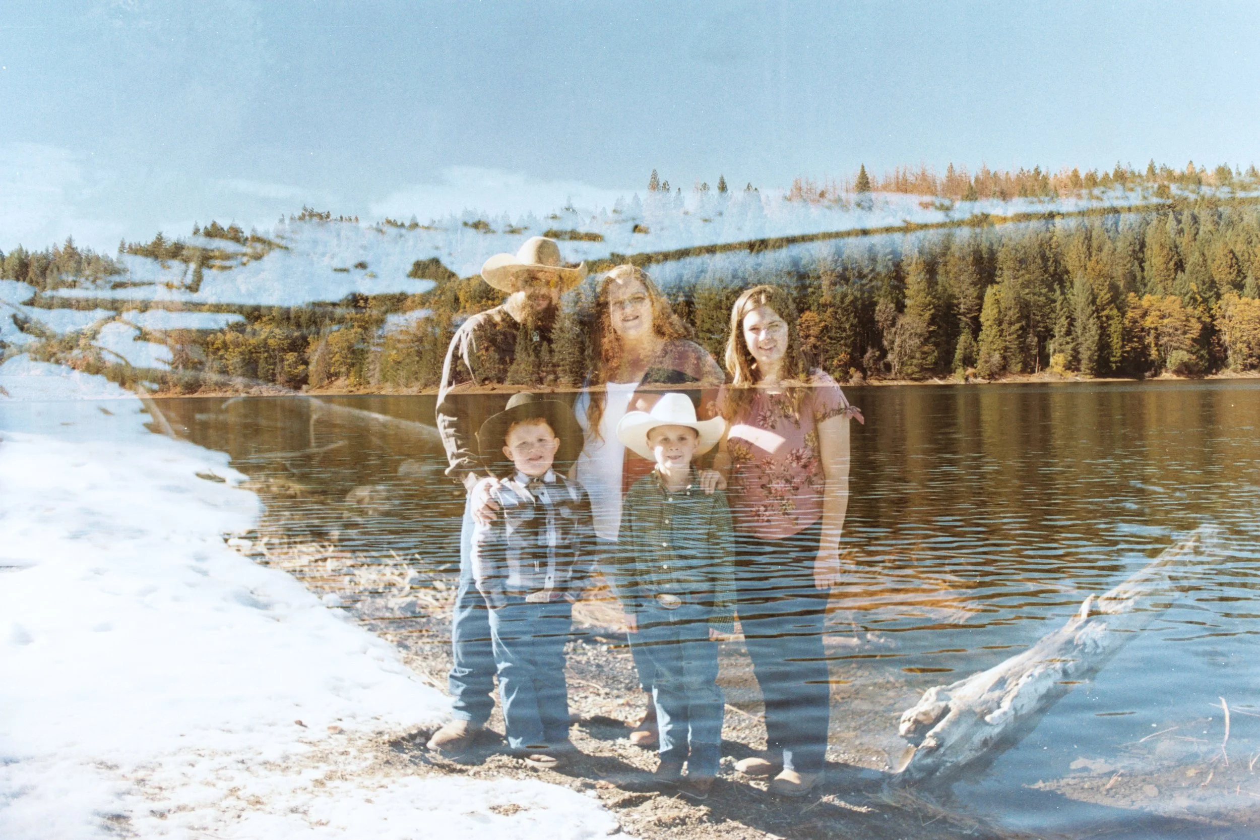 A family of five posing outdoors near a lake with a snowy forest and hills in the background, appearing to be a double exposure photo combining two images.