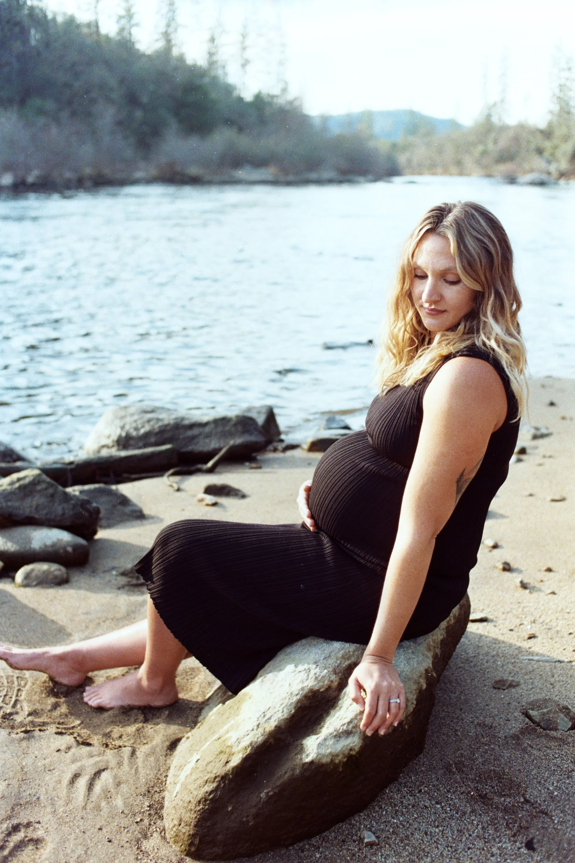 A pregnant woman in a black dress sitting on rocks at the beach, with the ocean and distant trees in the background.